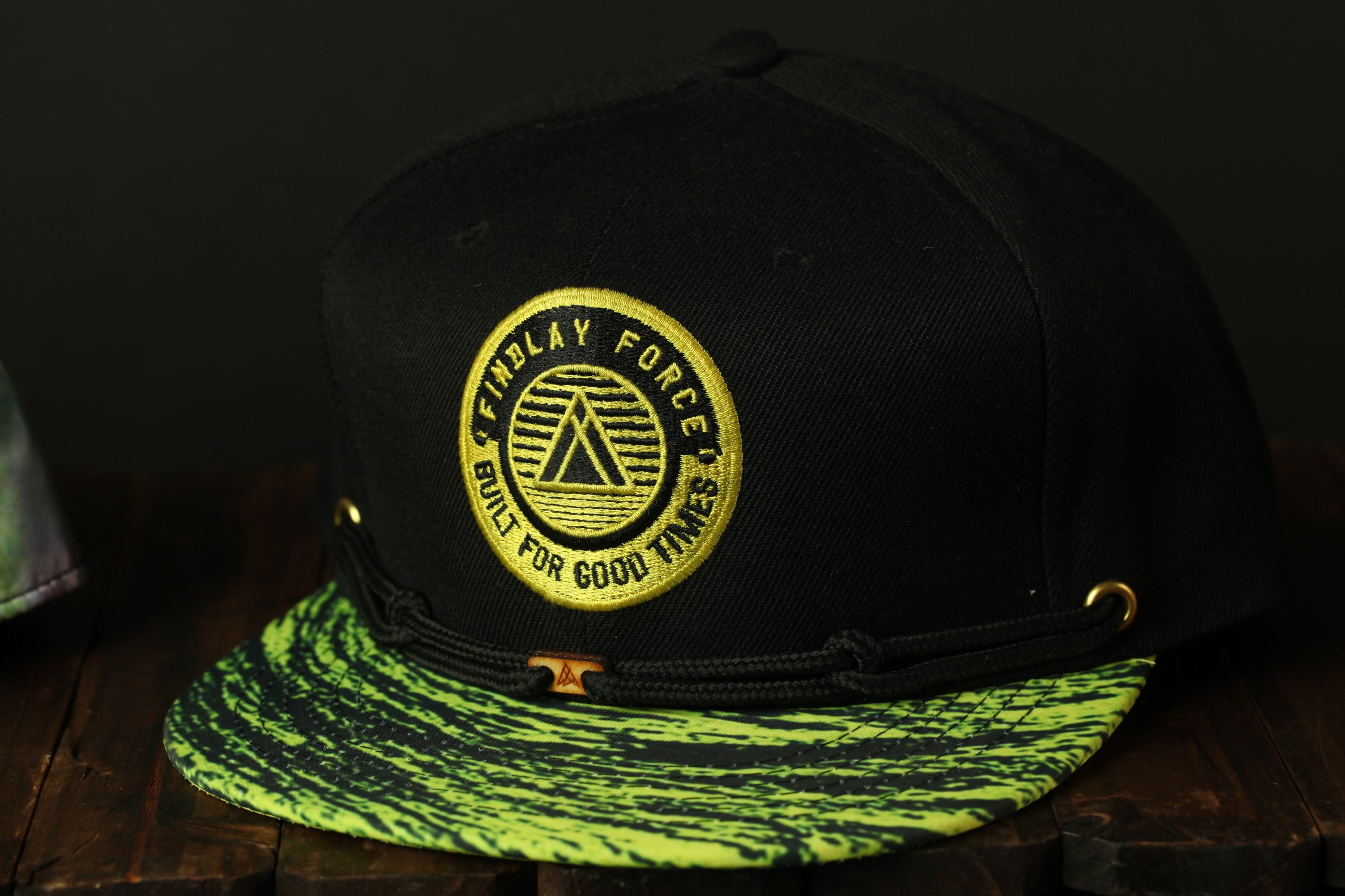 A black baseball cap with a yellow and green patterned brim, featuring a circular logo with text and a triangle symbol.