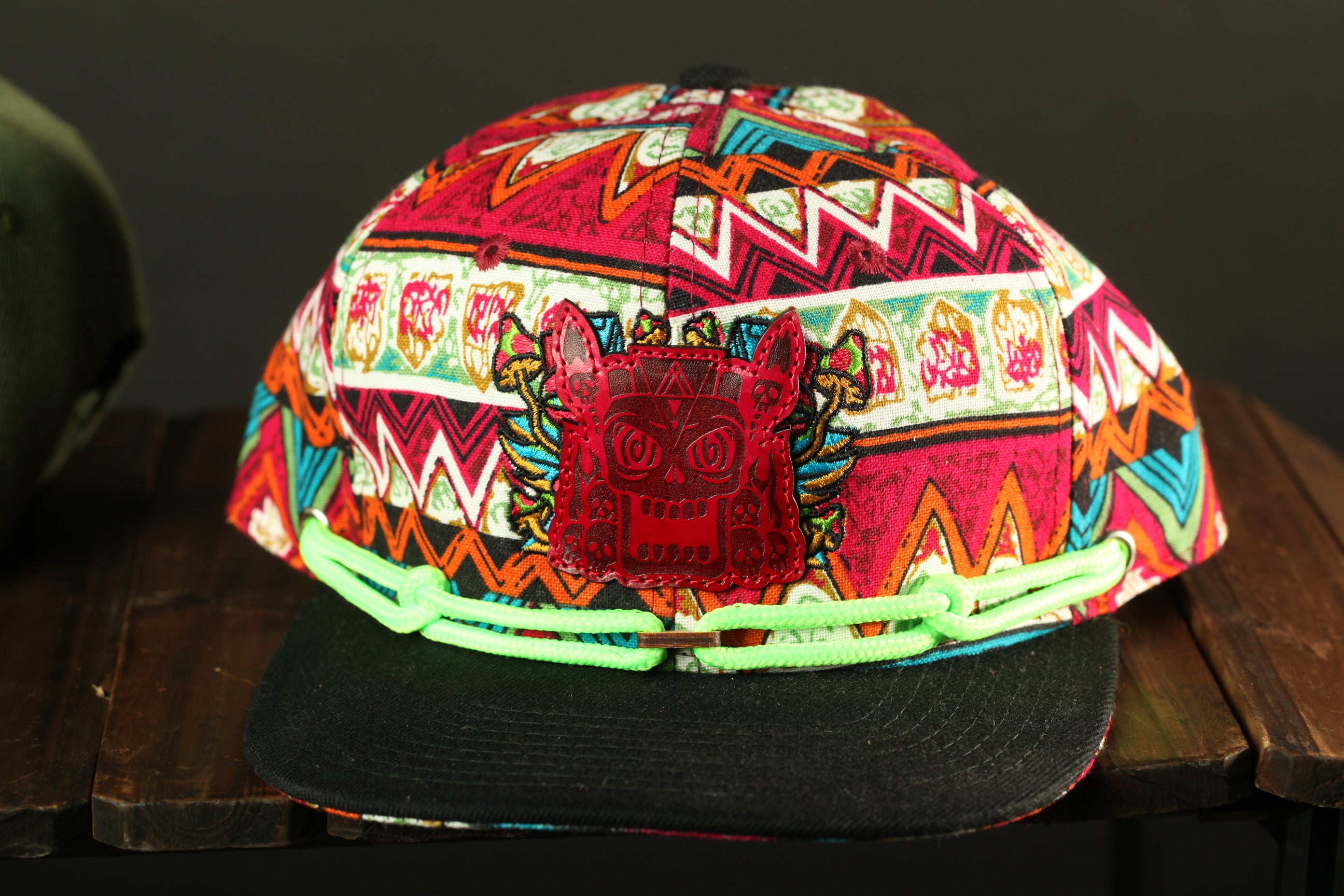A colorful, patterned baseball cap with a red skull-like logo on the front, displayed on a wooden surface.