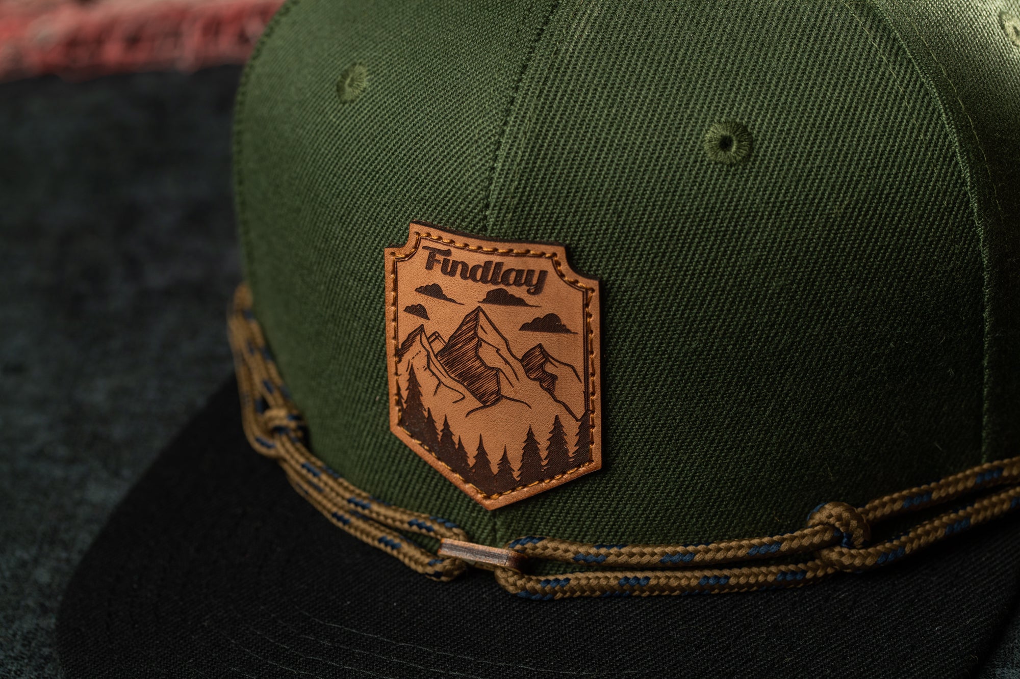 A close-up of a green baseball cap with a leather patch featuring a mountain landscape and the word "Findlay" printed on it.