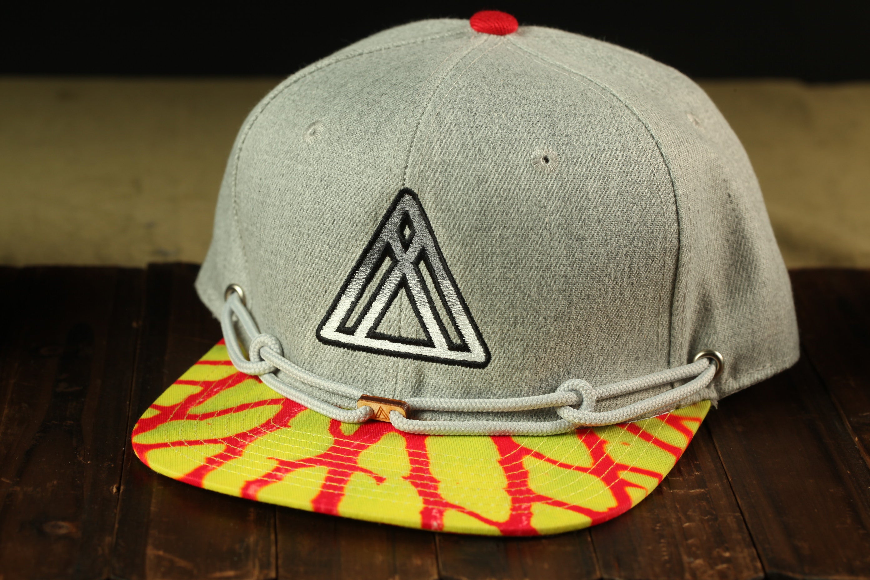 A gray baseball cap with a yellow and red patterned brim, featuring a black triangle logo on the front.