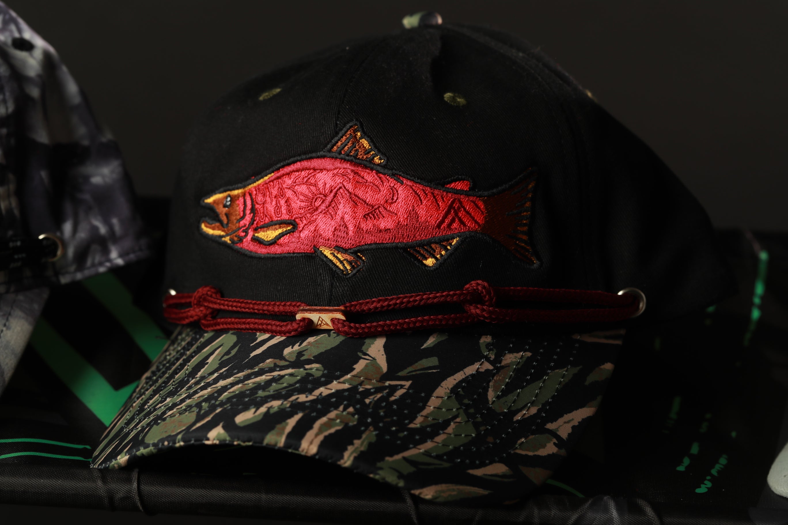 A black baseball cap with a red fish embroidered on it, featuring a camouflage pattern.