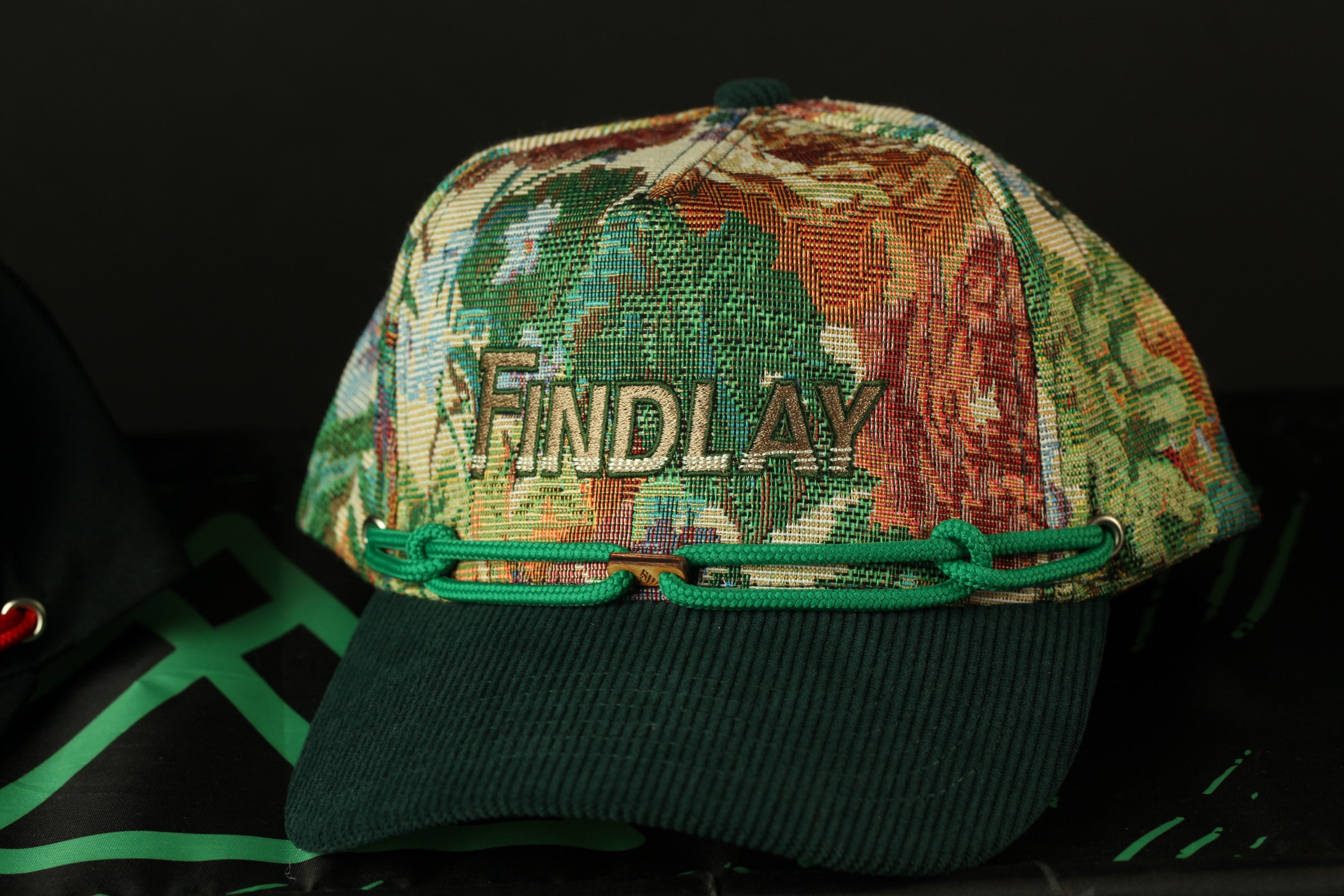 A colorful, patterned baseball cap with the word "FINDLAY" printed on it, sitting on a dark surface.