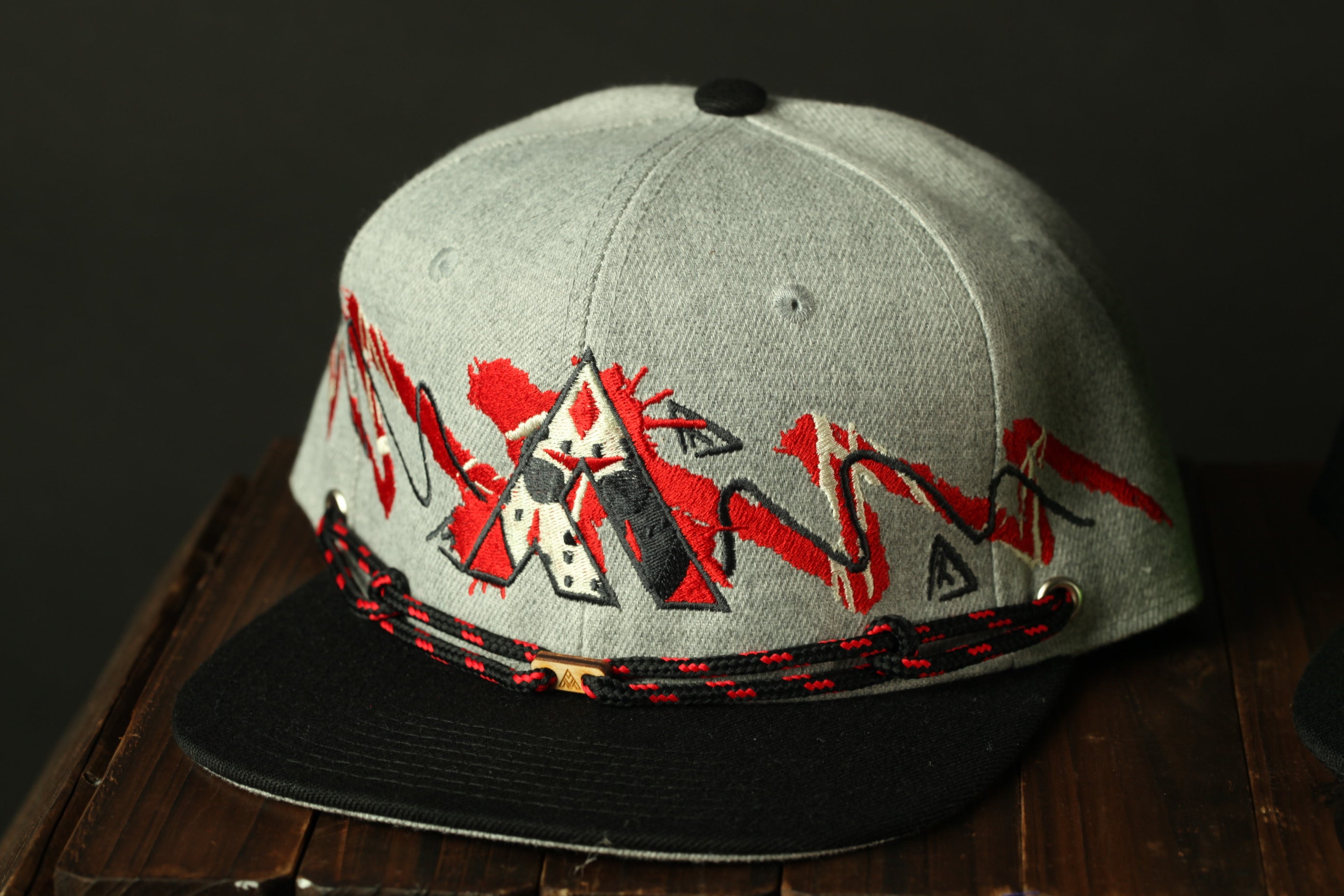 A gray baseball cap with red and black geometric designs on the front, sitting on a wooden surface.