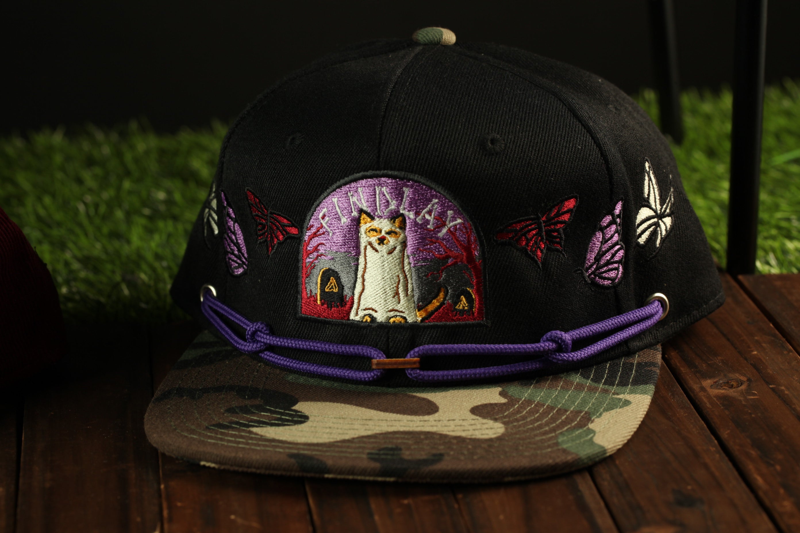 A black baseball cap with a camouflage pattern and a cat embroidered on it, sitting on a wooden surface.