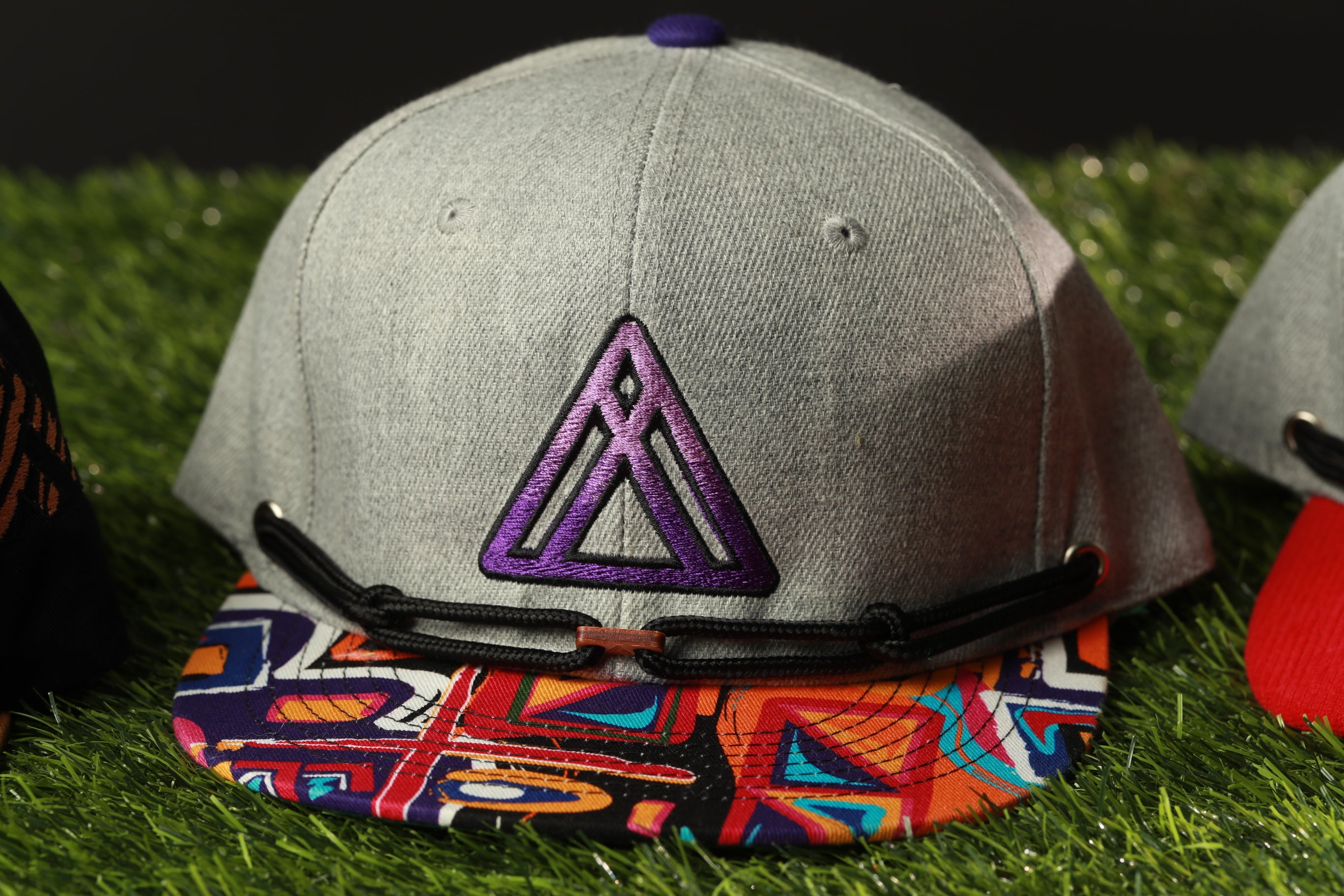 A gray baseball cap with a purple triangle logo and a colorful patterned fabric band, placed on a grassy surface.