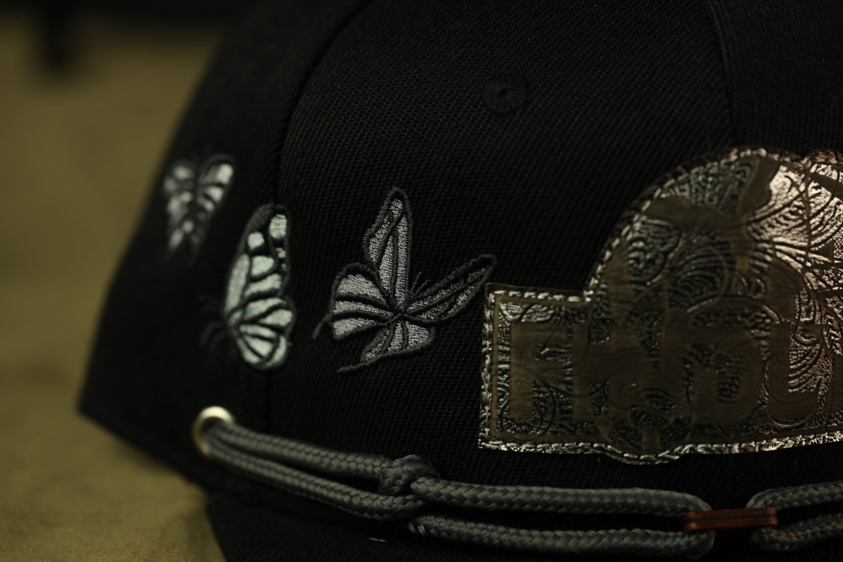 A close-up view of a black baseball cap with silver embroidered butterflies and a metal patch on the front.