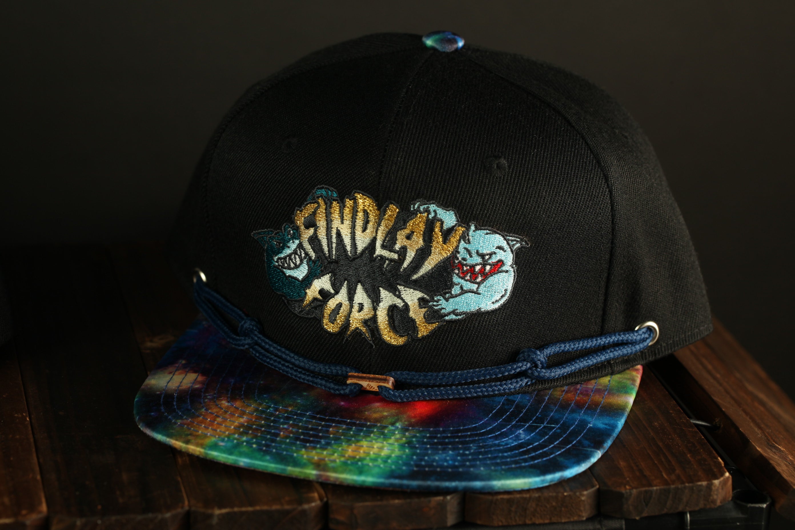 A black baseball cap with a tie-dye pattern and the text "Findlay Force" printed on it, sitting on a wooden surface.