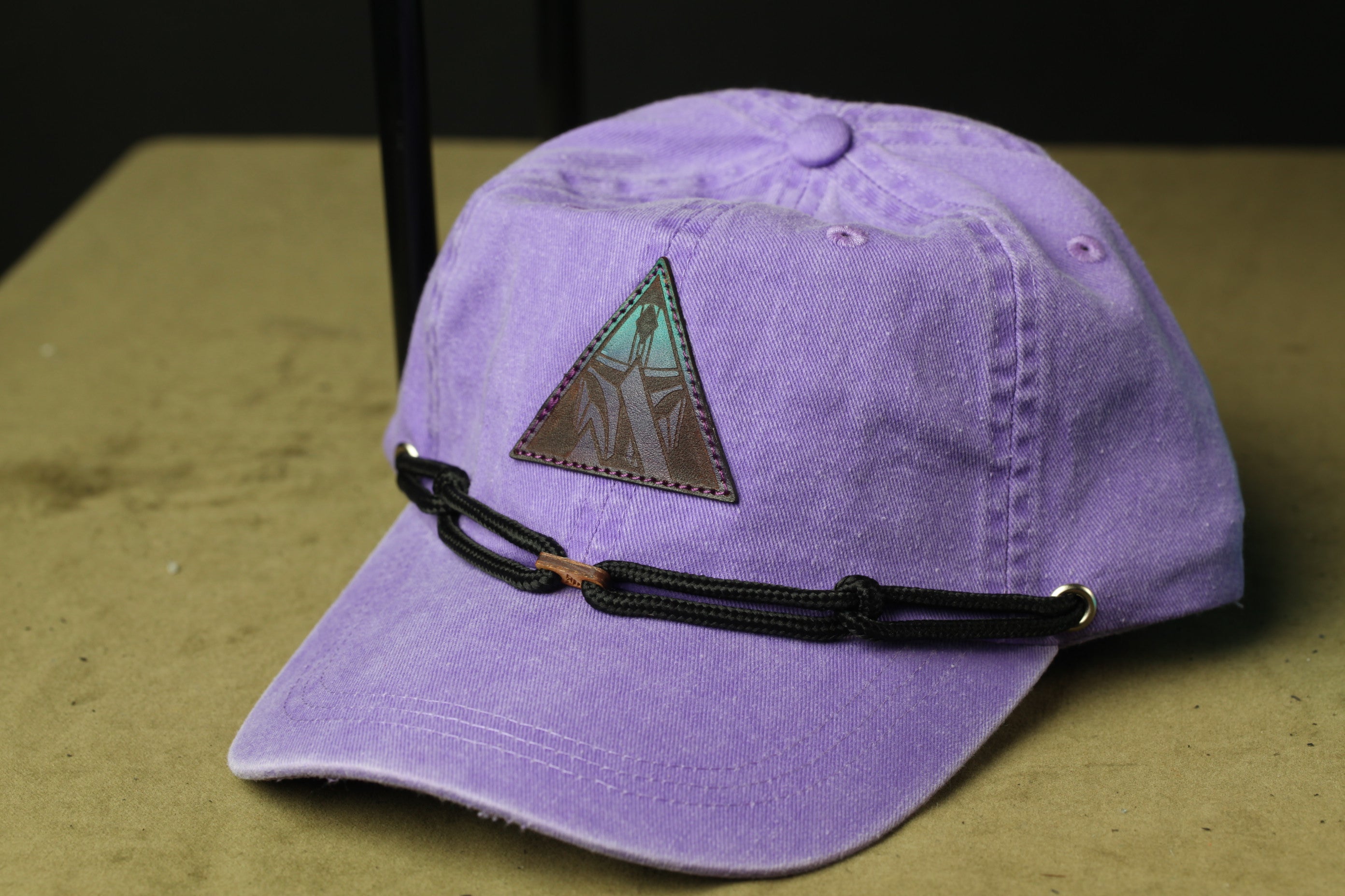 A purple baseball cap with a triangular patch on the front, featuring a design, is shown on a wooden surface.