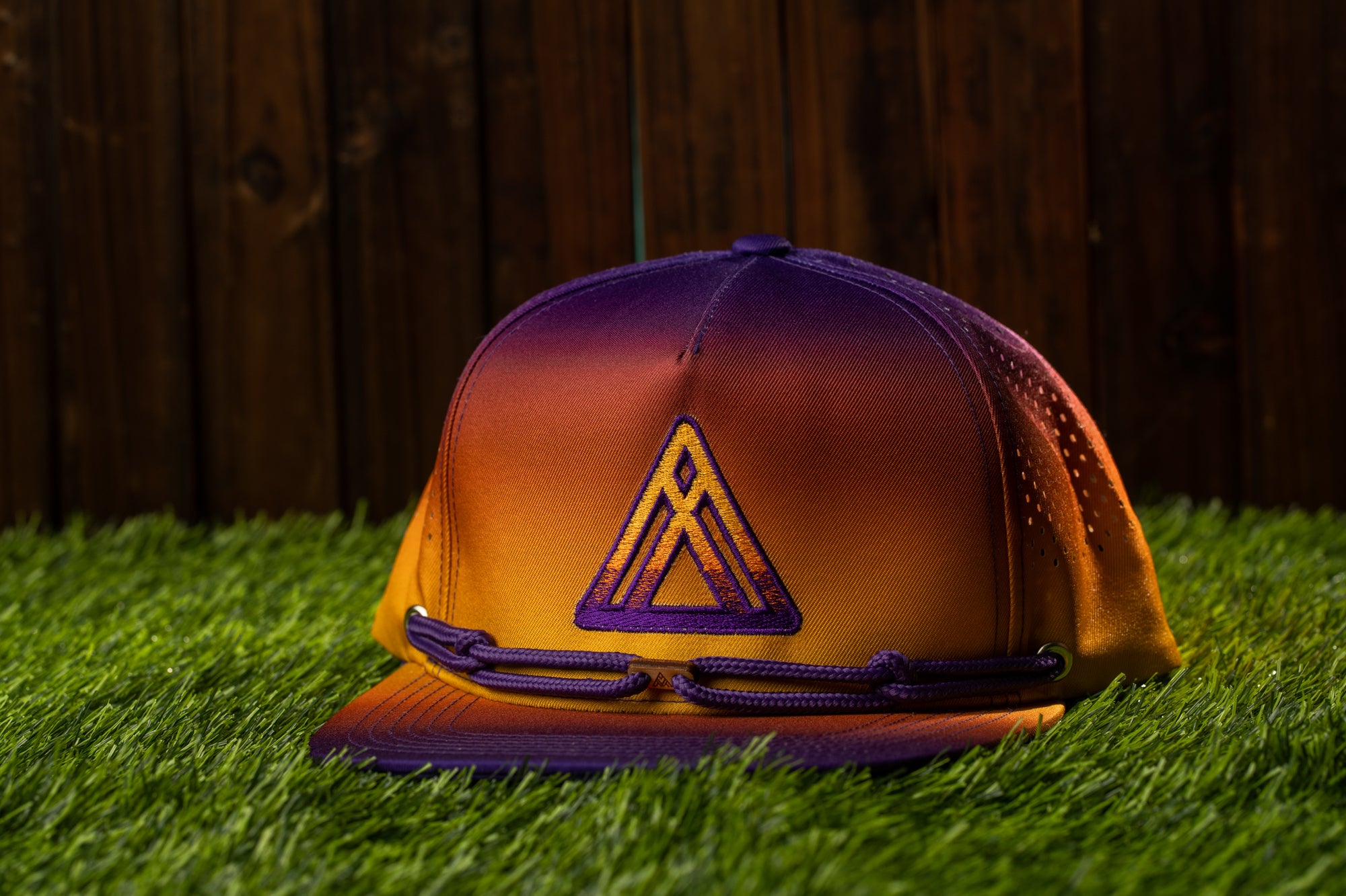 A purple and orange baseball cap with a triangular logo is placed on a grassy surface against a wooden fence background.
