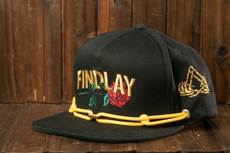 A black baseball cap with gold embroidered text and a red rose design, placed on a wooden surface.
