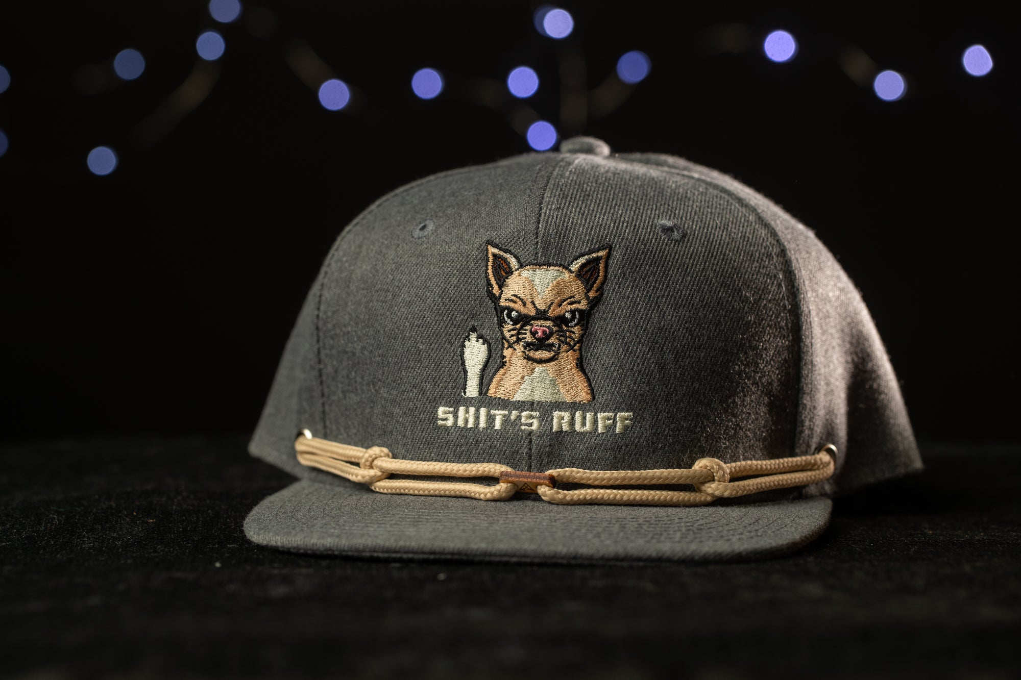 A gray baseball cap with a cartoon dog and the text "SHIT'S RUFF" printed on it, against a dark background with blue lights.