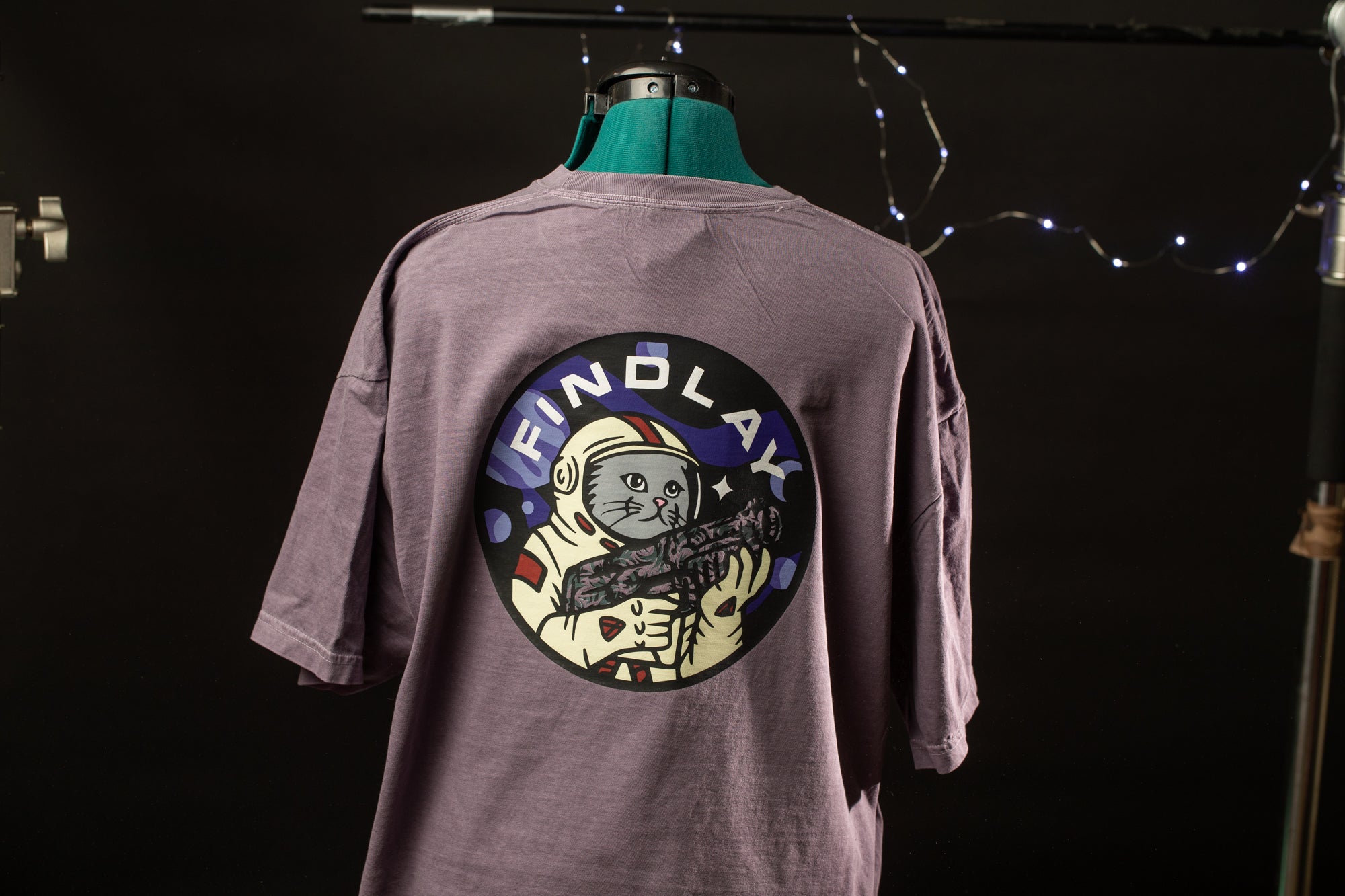 A purple t-shirt with a circular graphic design featuring a cat wearing a spacesuit and holding a rocket ship, with the text "FIND LAY" printed on the back.