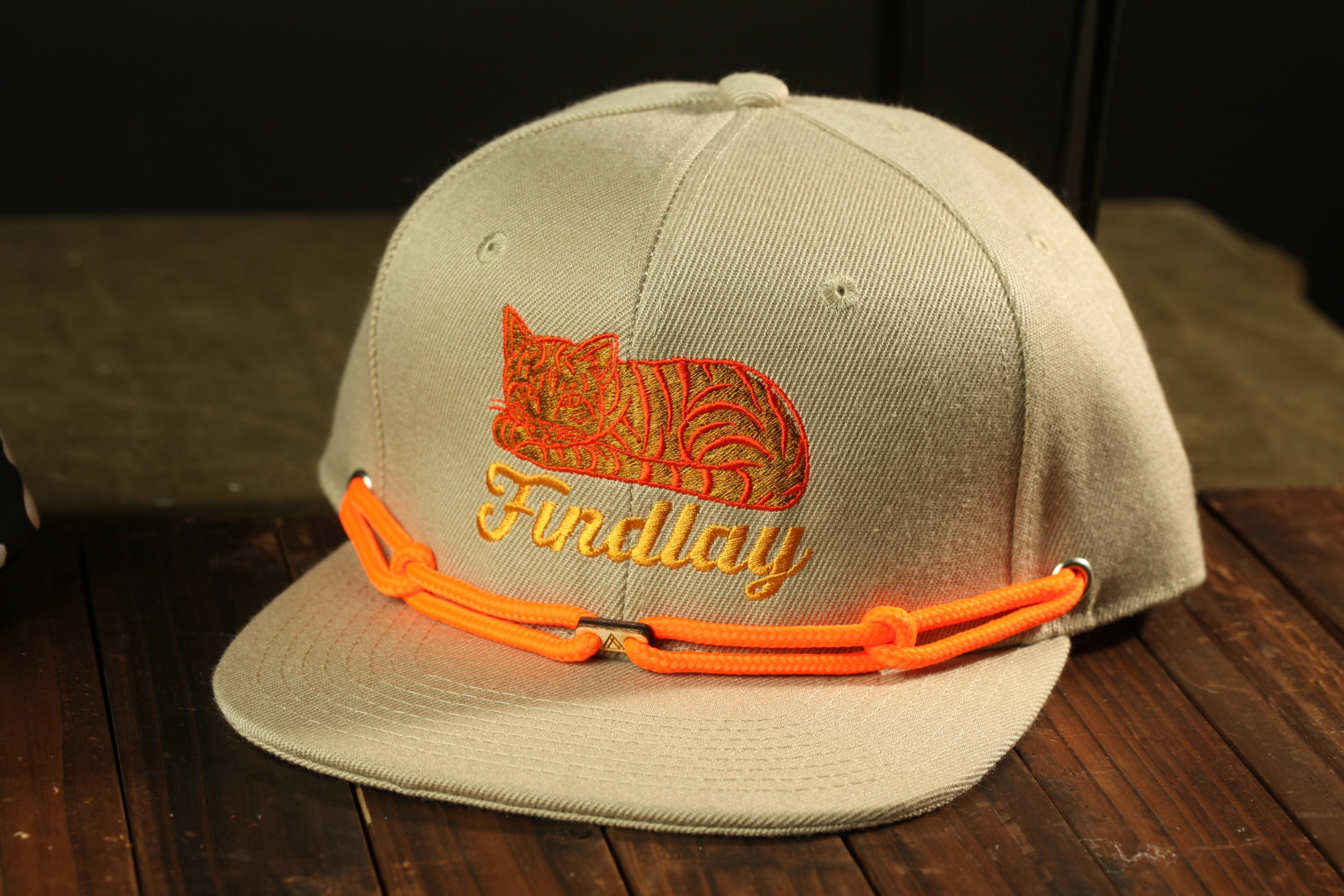 A beige baseball cap with an orange cat embroidered on it, sitting on a wooden surface.
