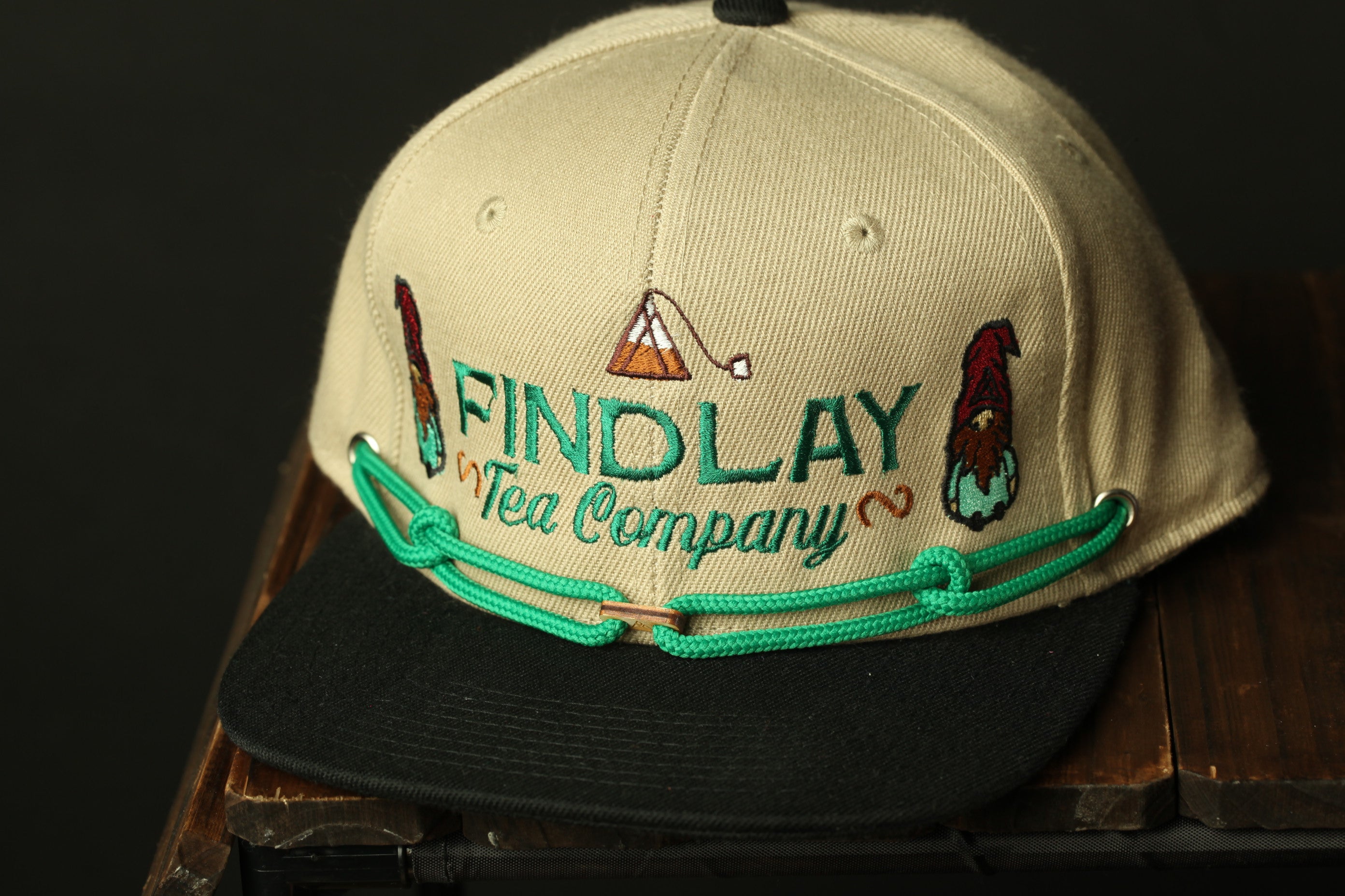 A beige baseball cap with the text "FIND LAY Tea Company" embroidered on it, along with two small gnome-like figures on either side of the text.