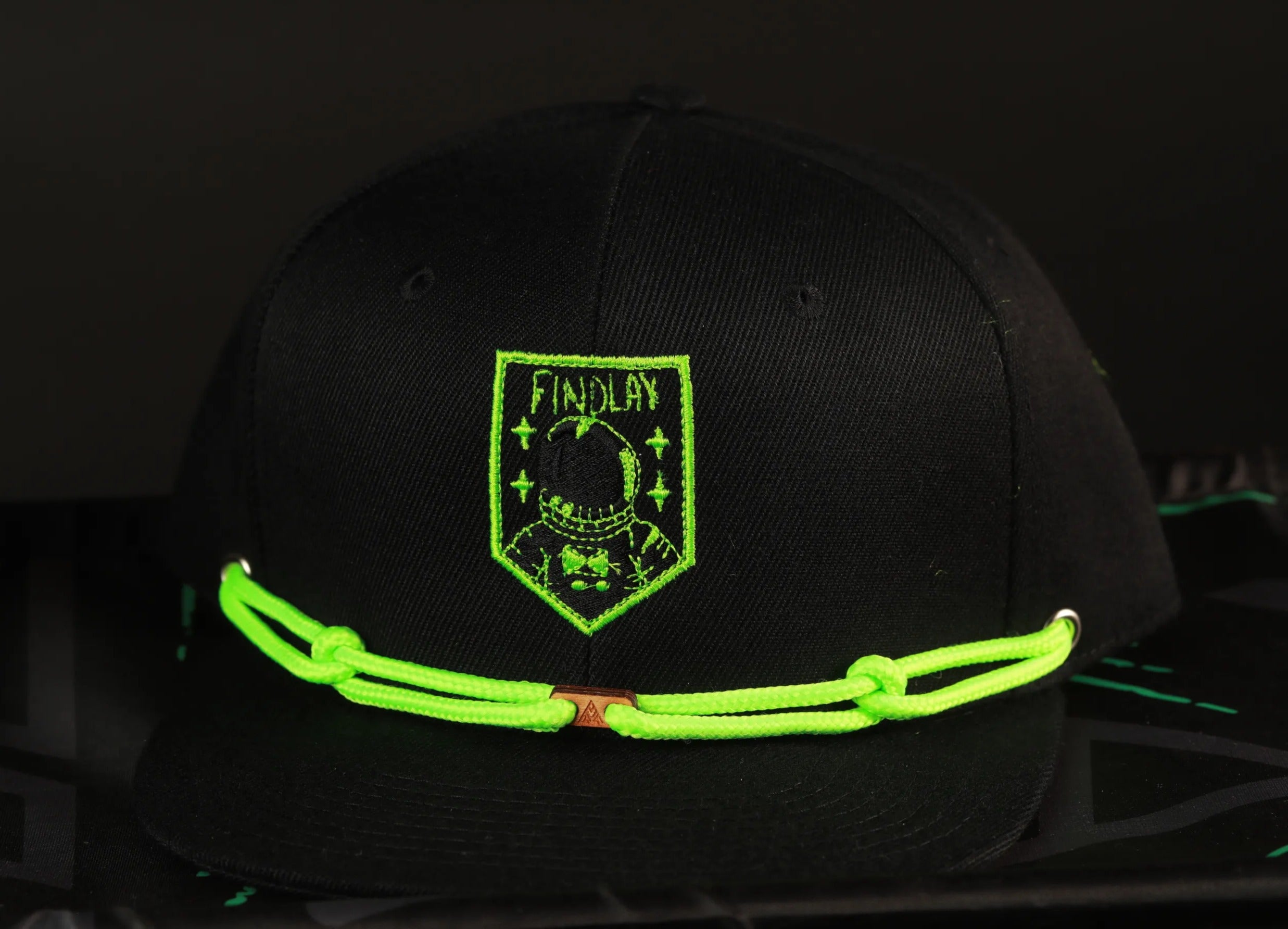 A black baseball cap with a green embroidered patch on the front, featuring a skull and crossbones design.