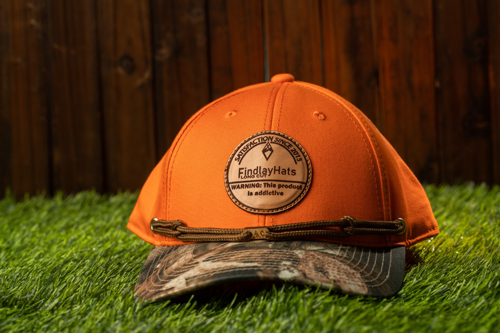 An orange baseball cap with a camouflage pattern and a circular logo on the front, placed on a grassy surface.