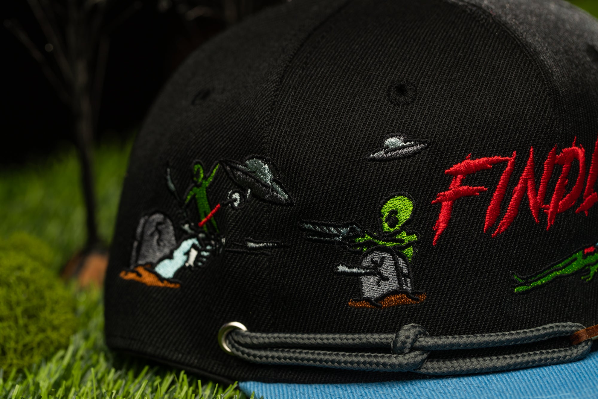 a black baseball cap with colorful embroidered designs on it, including a scene with a green alien and a human character.