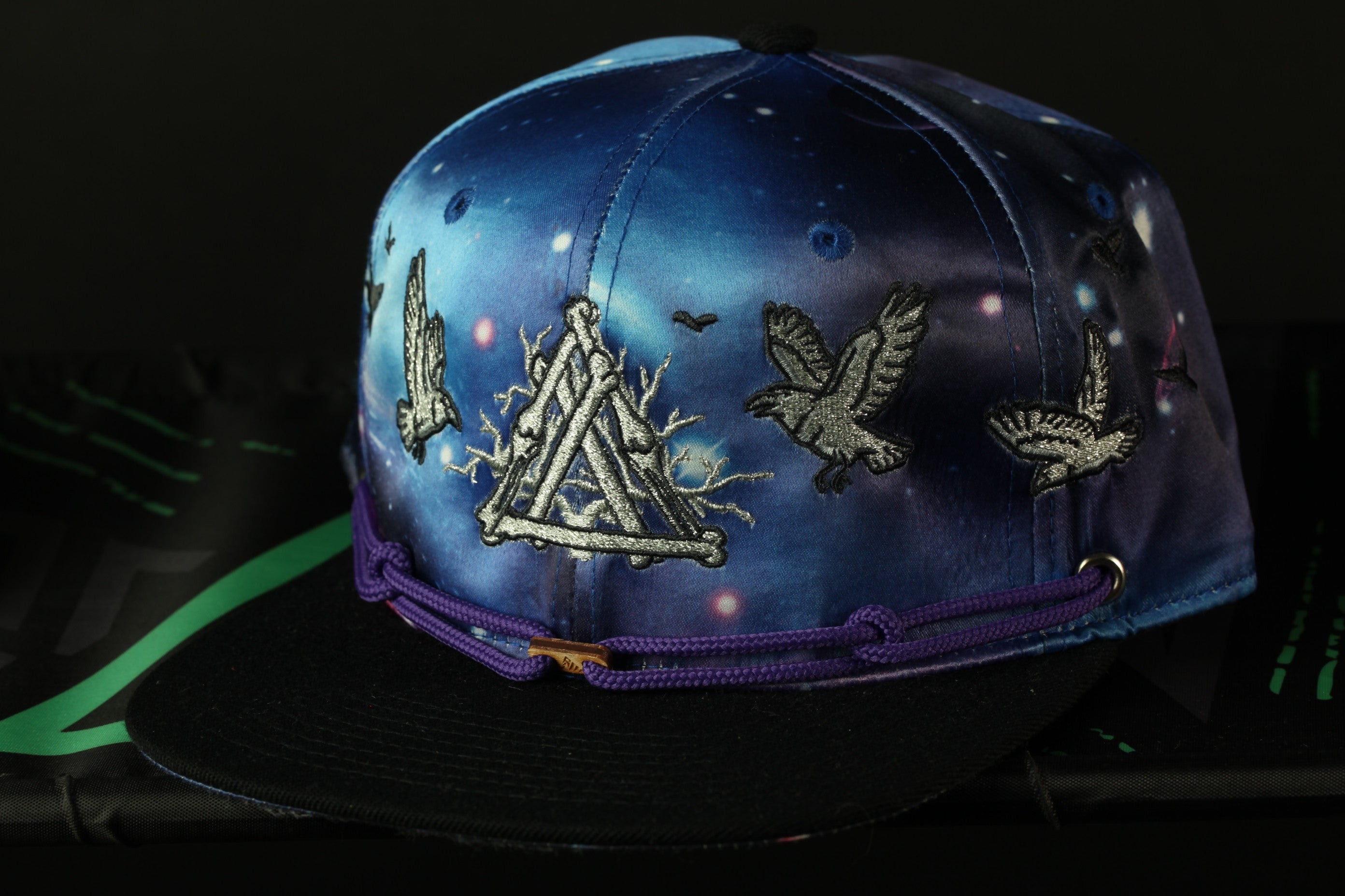 A blue baseball cap with a purple band and a design featuring a triangle, birds, and stars against a night sky background.
