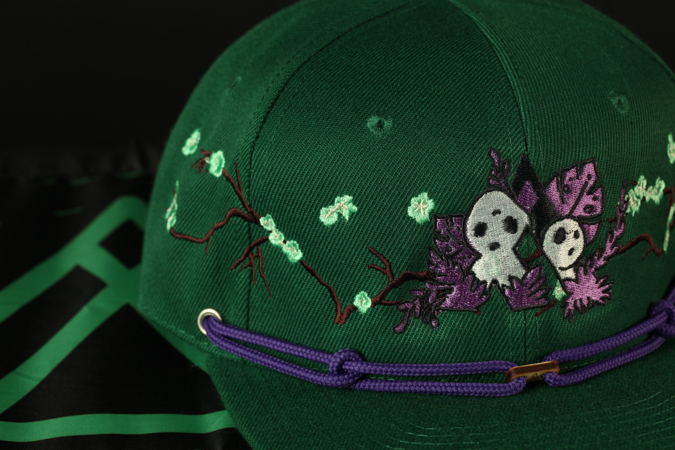 A close-up view of a green baseball cap with purple and white floral embroidery, featuring a small skull design.