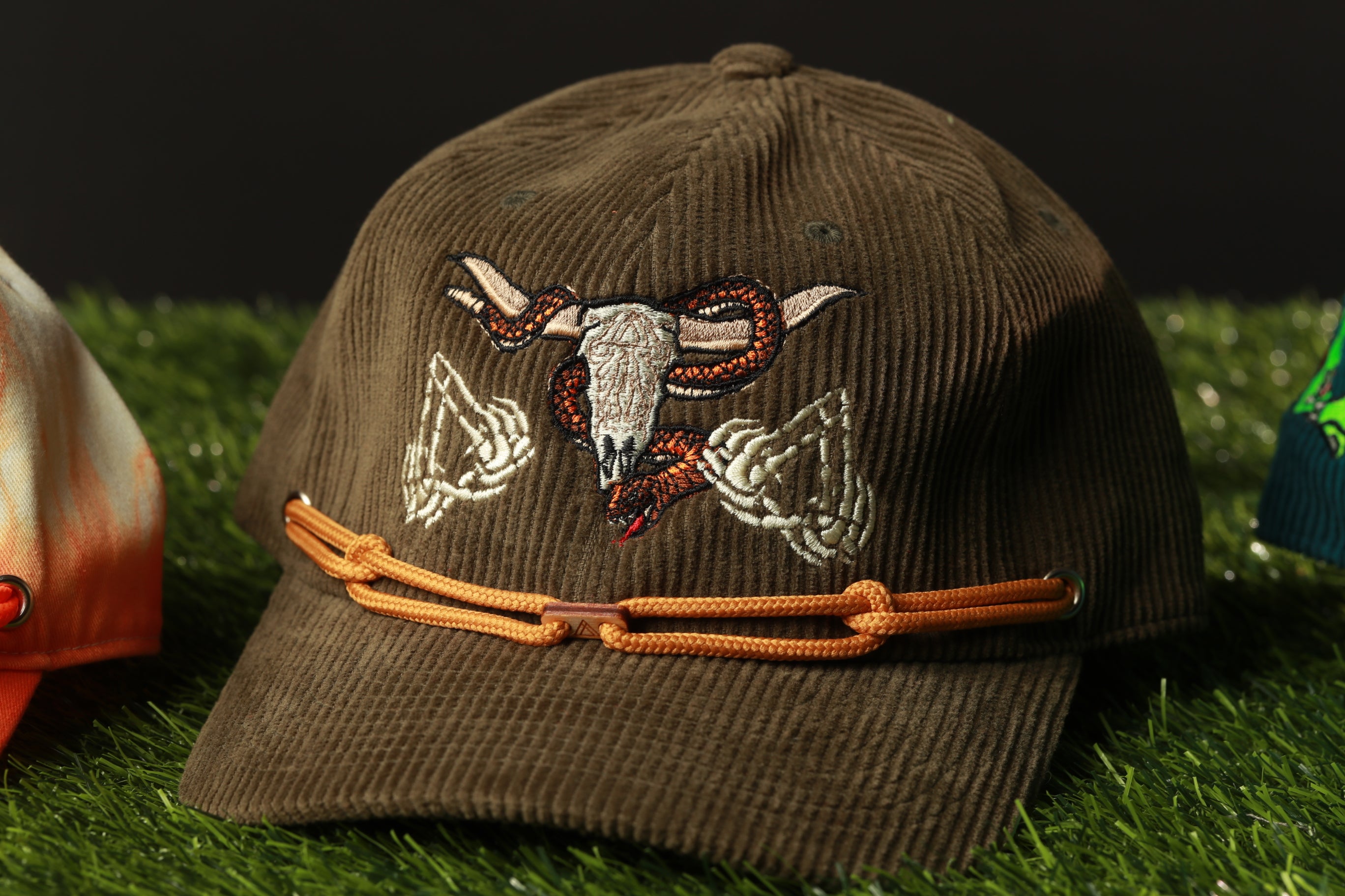 A brown baseball cap with an embroidered design of a skull and snake on the front, sitting on a grassy surface.