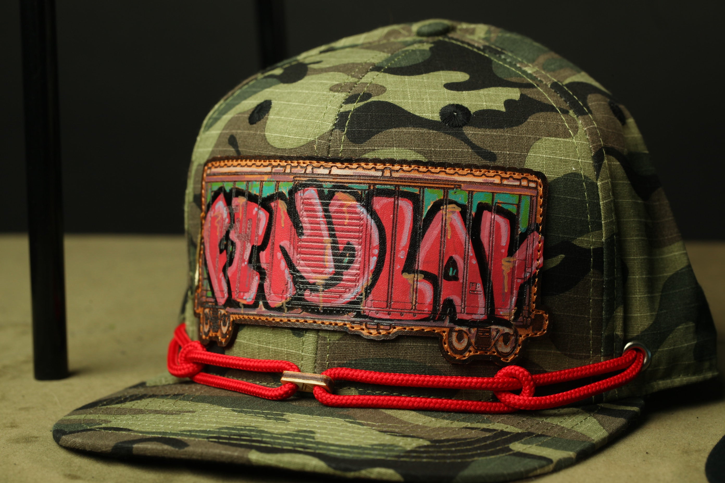 A camouflage baseball cap with a red chain and a patch that says "FIND LAY" in graffiti-style lettering.