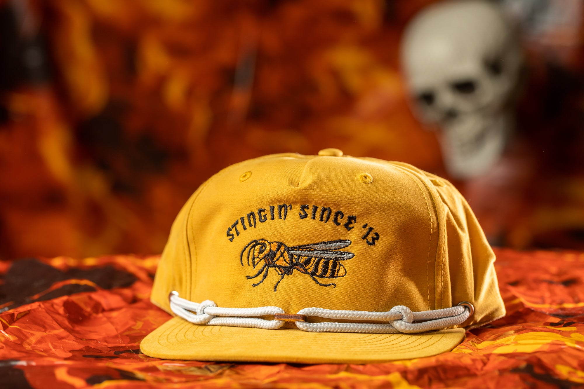 A yellow baseball cap with a graphic of a bee and the text "Stingin' since 13" is displayed on a red and orange patterned surface.