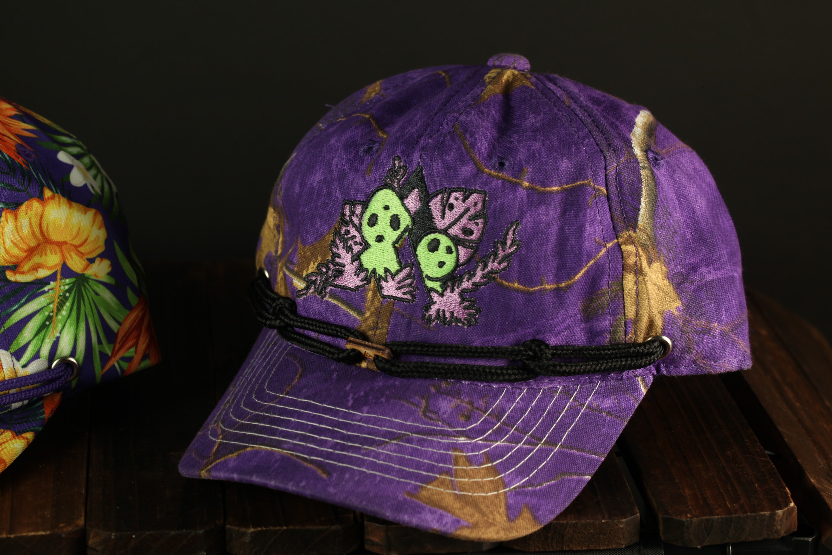 A purple baseball cap with a floral pattern and a green skull design is displayed on a wooden surface.