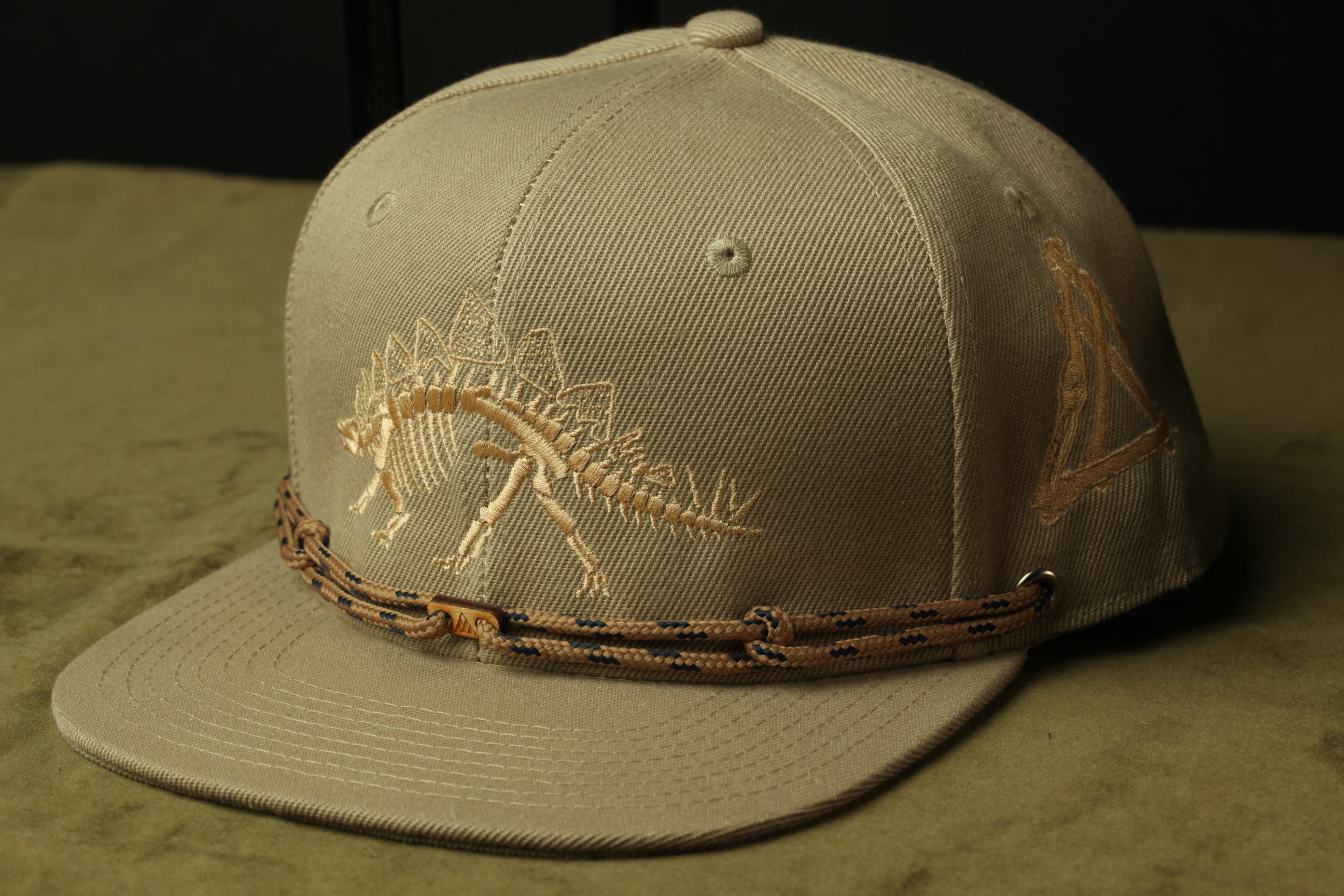 A beige baseball cap with a dinosaur skeleton embroidered on it, featuring a rope-like strap around the brim.