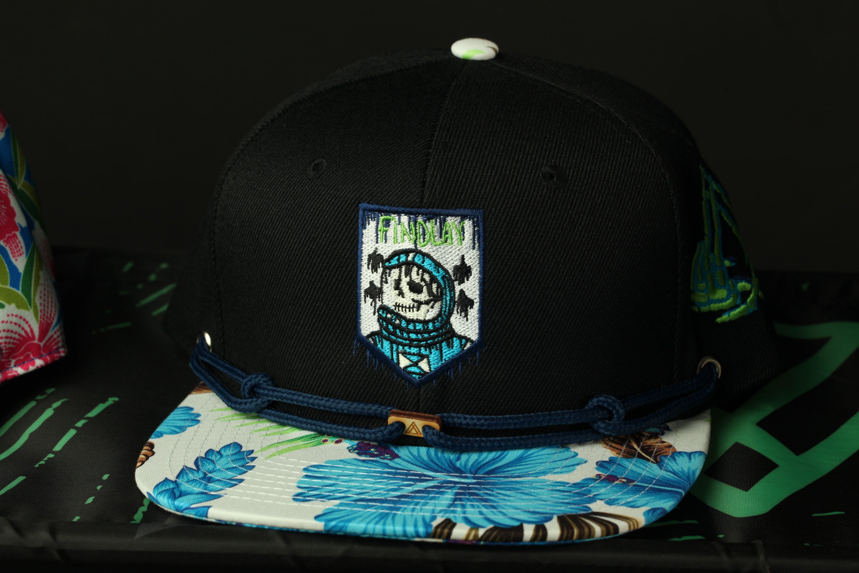 A black baseball cap with a floral pattern and a patch on the front, placed on a surface with a green and black design.