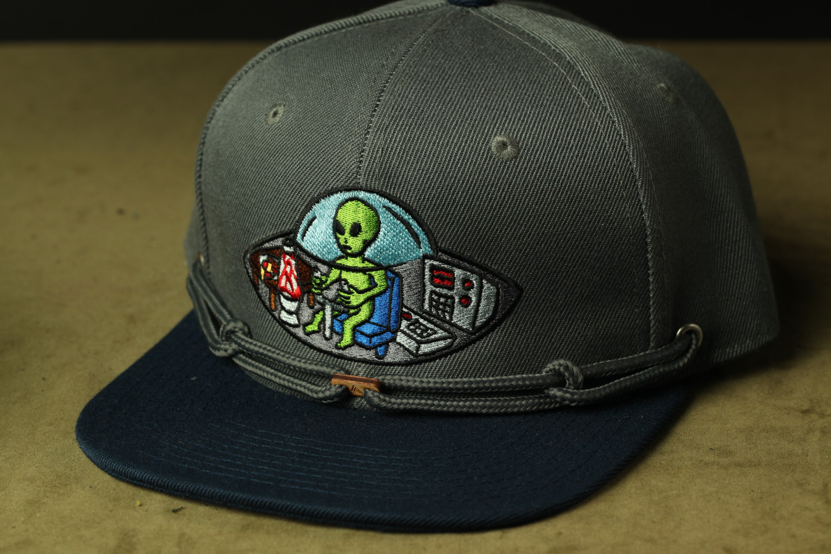 A gray baseball cap with a colorful embroidered design depicting an alien in a spaceship cockpit.