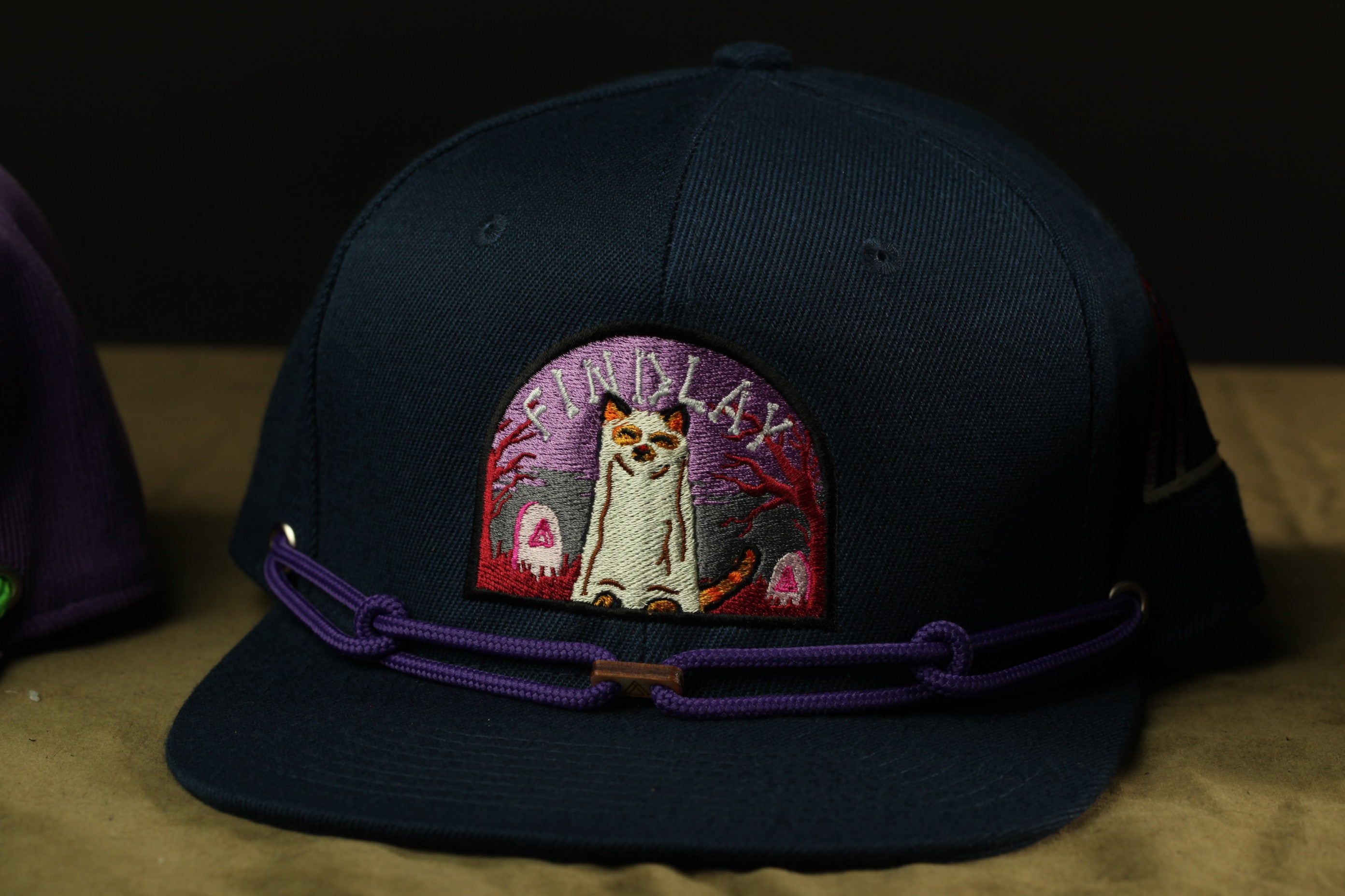 A dark-colored baseball cap with a purple string and a patch featuring a cat and the word "FLINDA" on it.