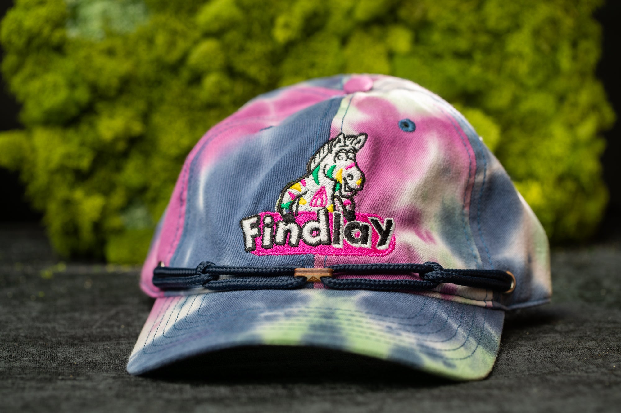 A tie-dye baseball cap with a colorful pattern and the word "Findlay" printed on it, placed on a surface with a green mossy background.