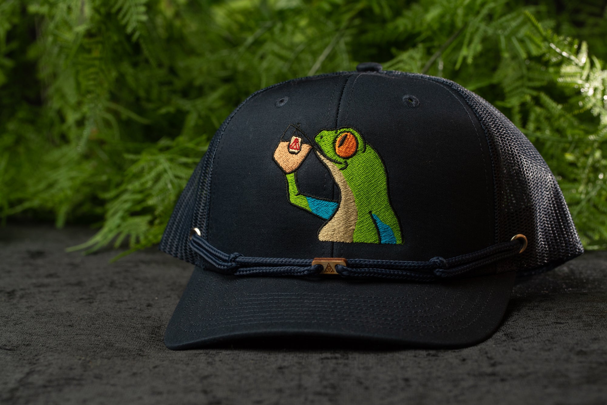 A black baseball cap with a colorful frog embroidered on it, placed on a surface with greenery in the background.
