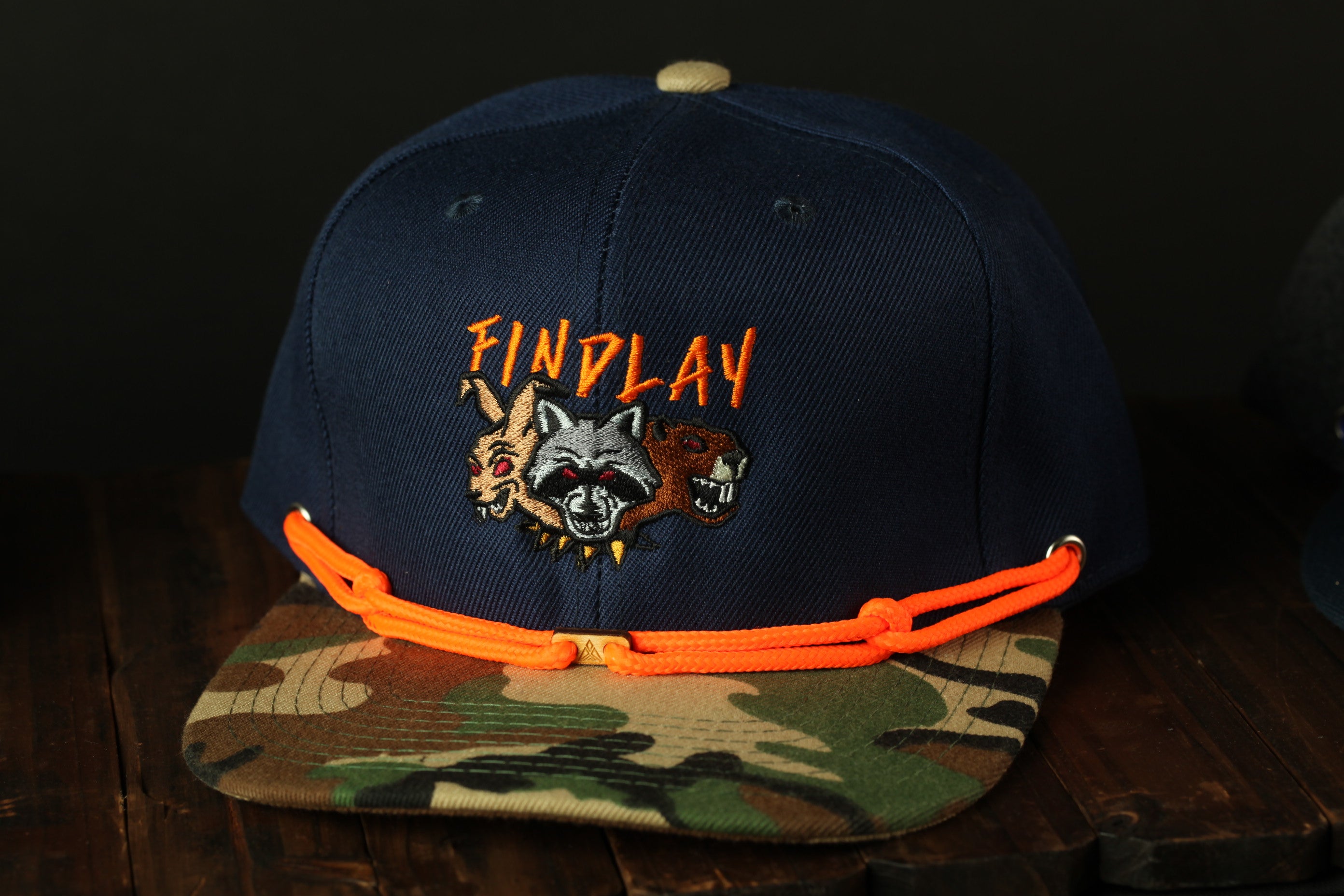 A navy blue baseball cap with a camouflage pattern and orange straps, featuring a graphic of a raccoon and the text "FINPLAY" on the front.