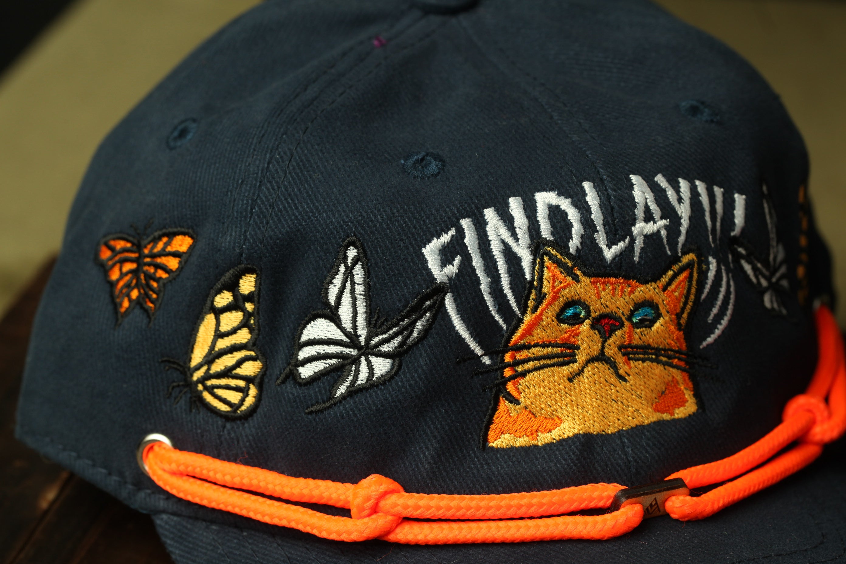 A close-up view of a black baseball cap with various embroidered designs, including a cat, butterflies, and a butterfly.