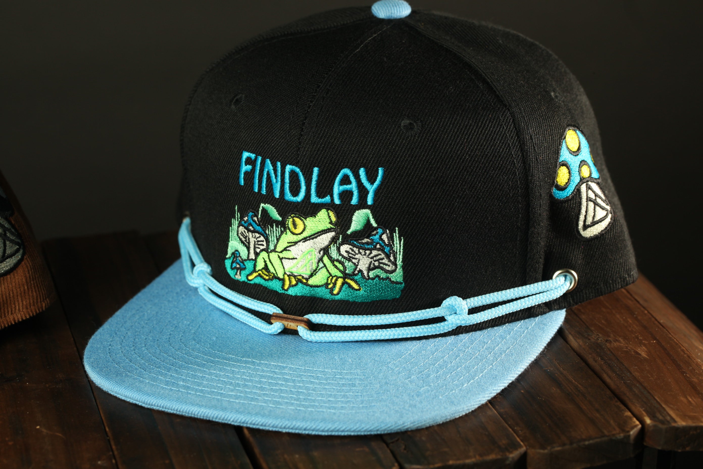 A black baseball cap with a blue brim and a graphic of a frog on the front, sitting on a wooden surface.