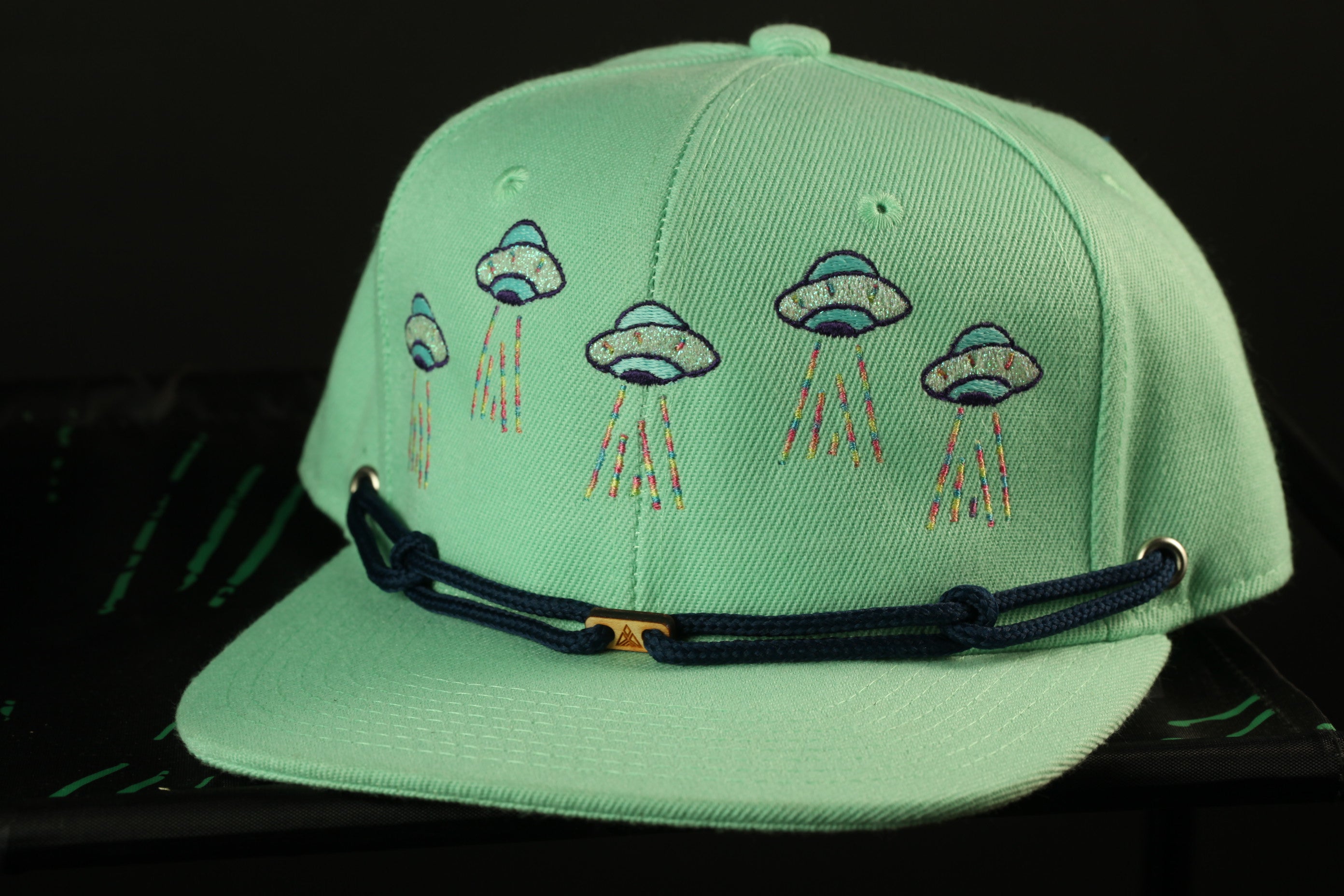 A mint green baseball cap with a black strap and embroidered designs of colorful alien spacecraft on the front.