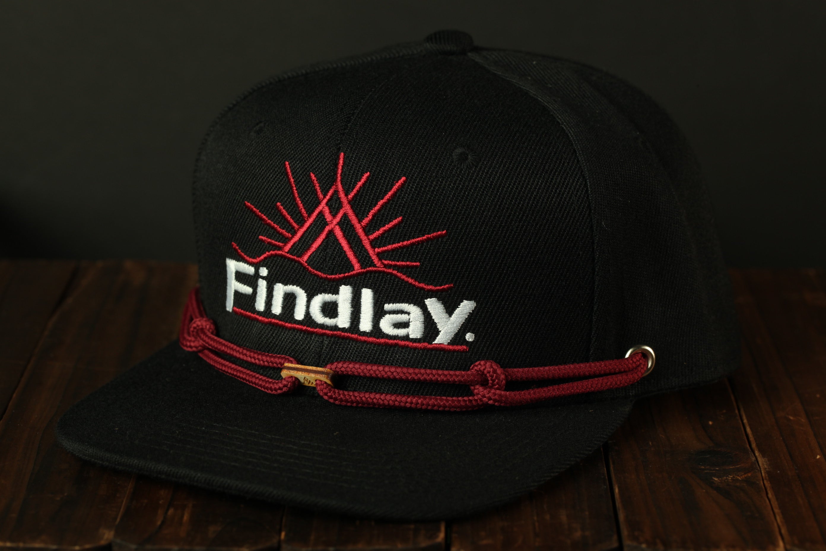 A black baseball cap with a red rope and the word "Findlay" printed on it, sitting on a wooden surface.