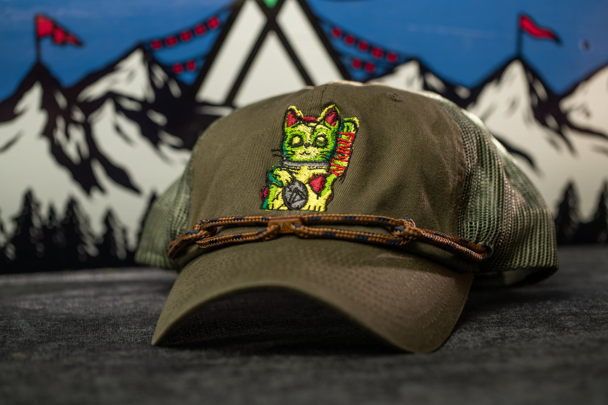 A green baseball cap with a colorful cat embroidered on it, placed on a gray surface against a background of mountains and flags.