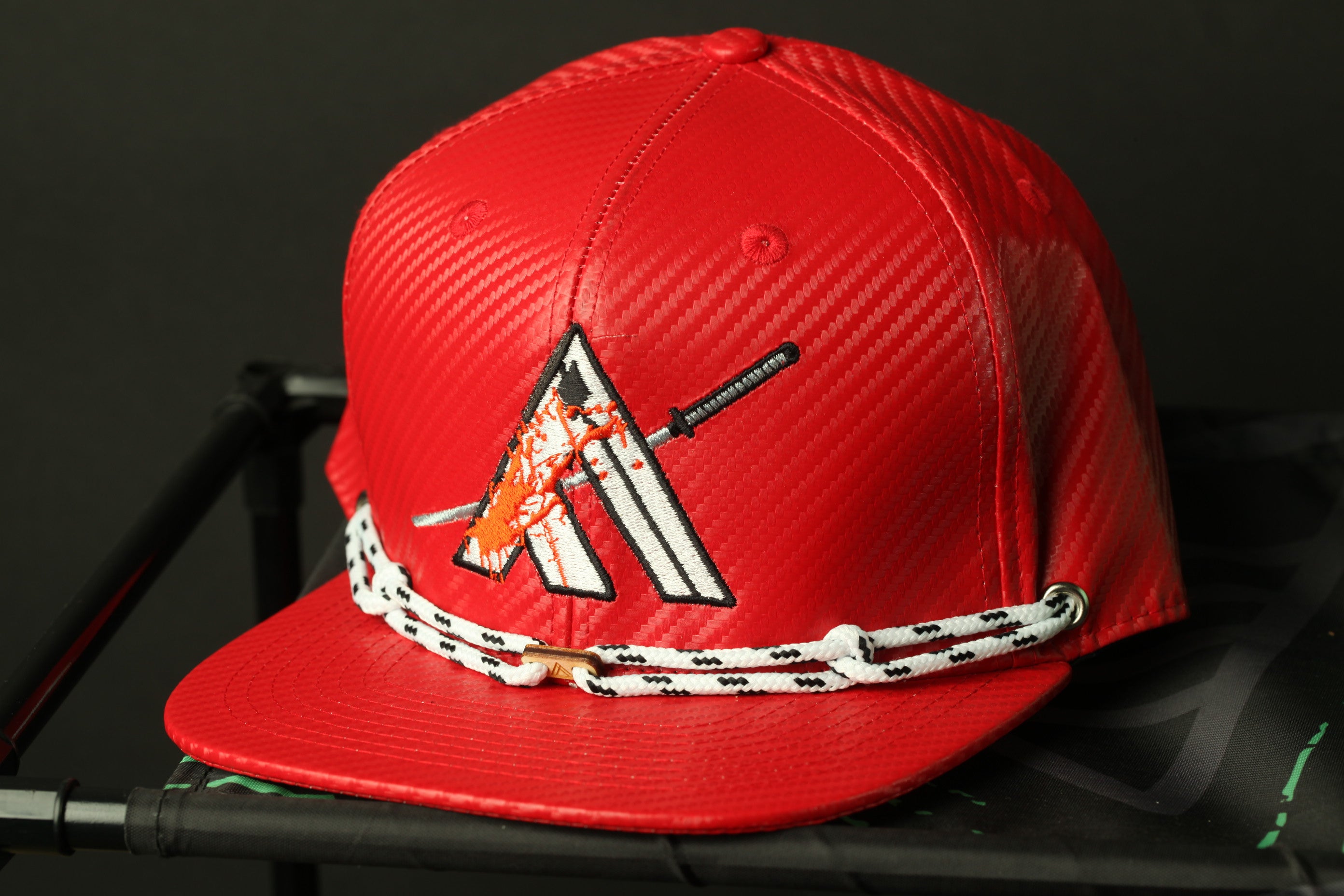 A red baseball cap with a white and orange design on the front, featuring a stylized letter 'A' and a sword-like emblem.