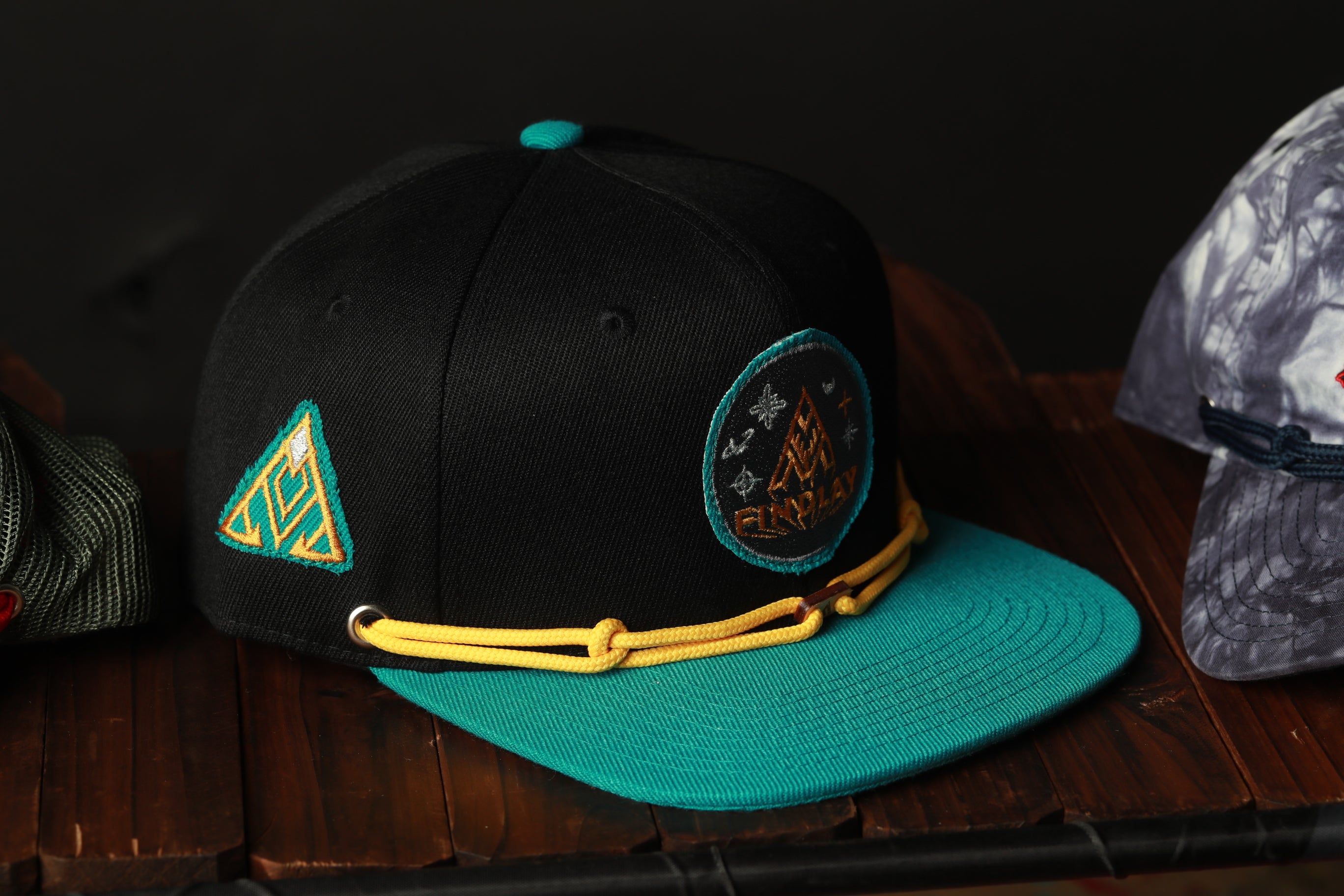 A black baseball cap with a teal brim and yellow string, featuring a circular patch with a graphic design, is displayed on a wooden surface.