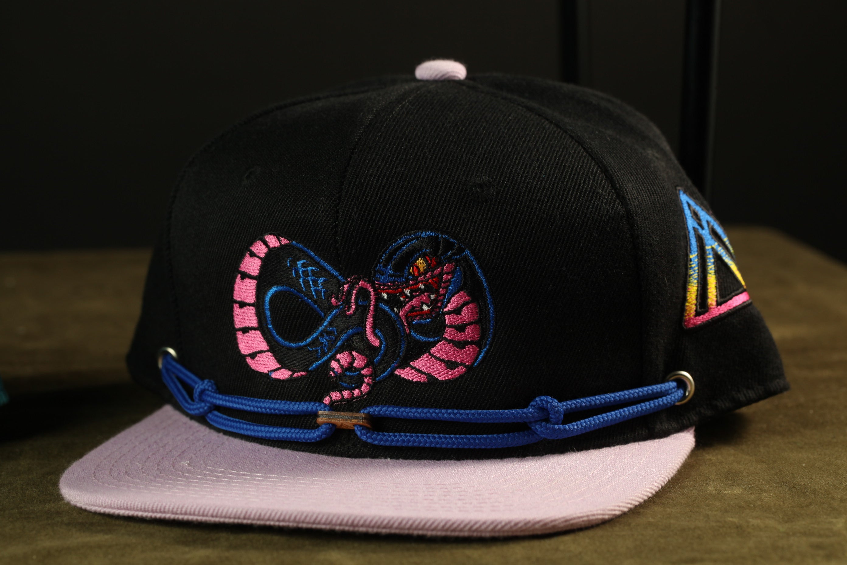 A black baseball cap with a colorful snake design on the front, featuring a pink and blue pattern.