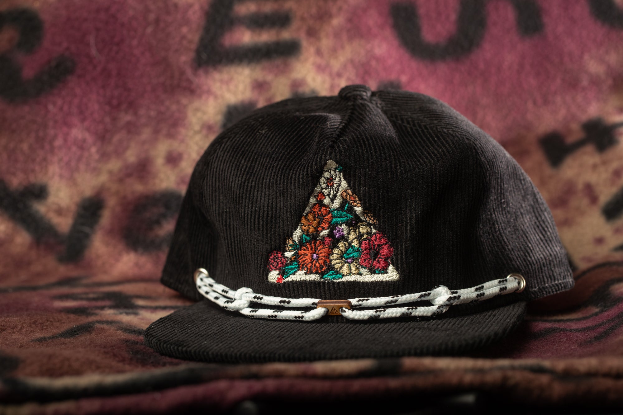 A black baseball cap with a colorful floral embroidered design on the front, resting on a patterned fabric background.