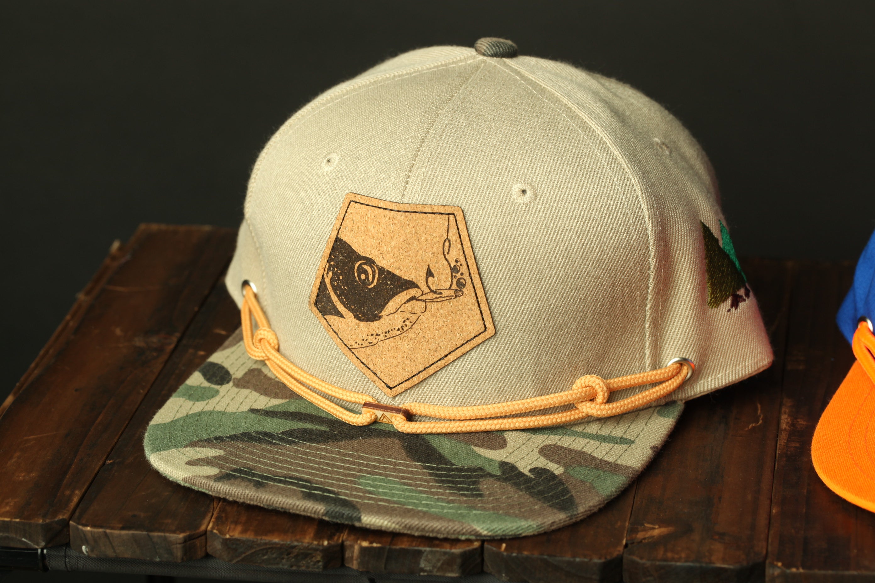 A beige baseball cap with a camouflage pattern and a fish-like patch on the front, sitting on a wooden surface.
