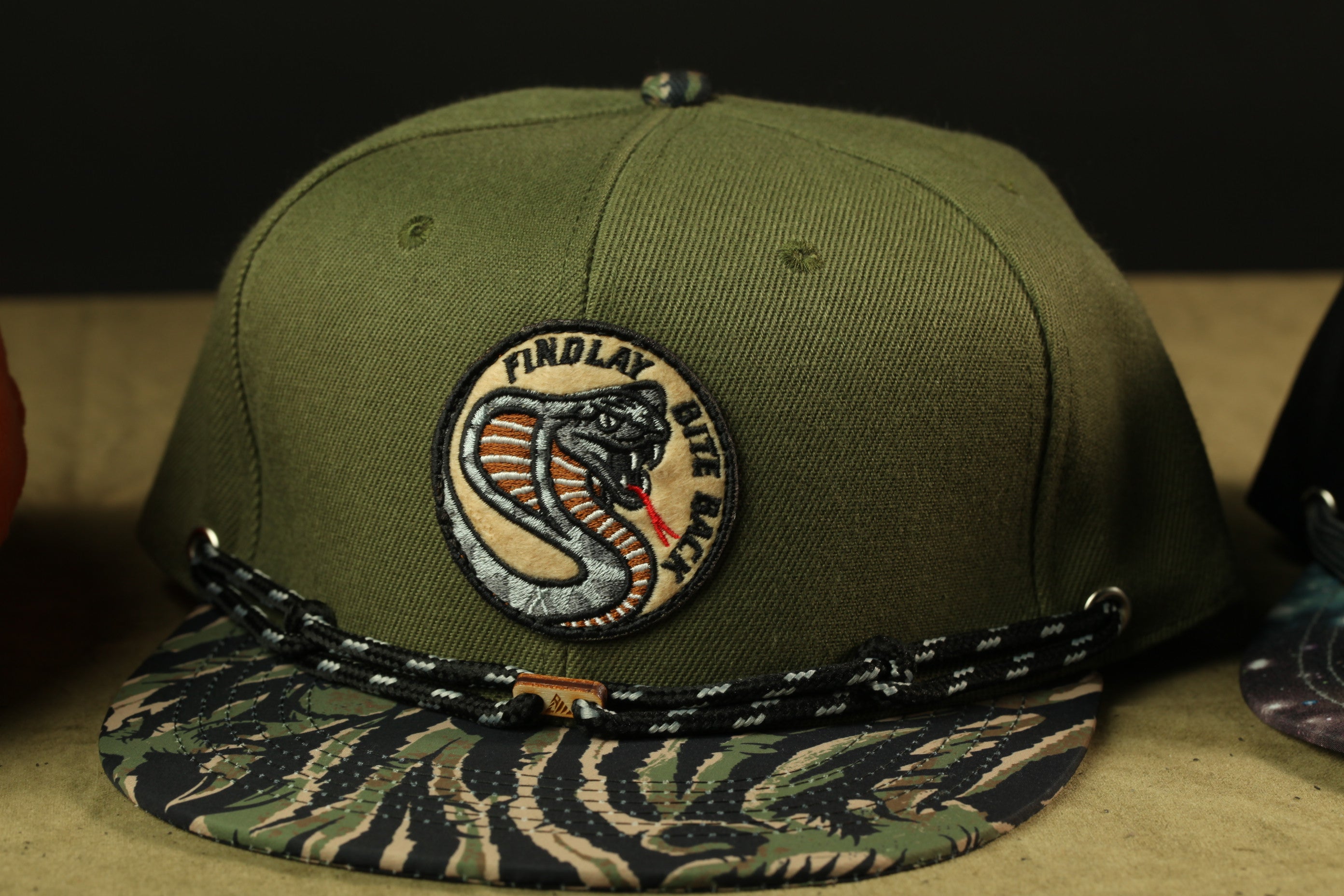 A green baseball cap with a camouflage pattern and a patch featuring a snake design.