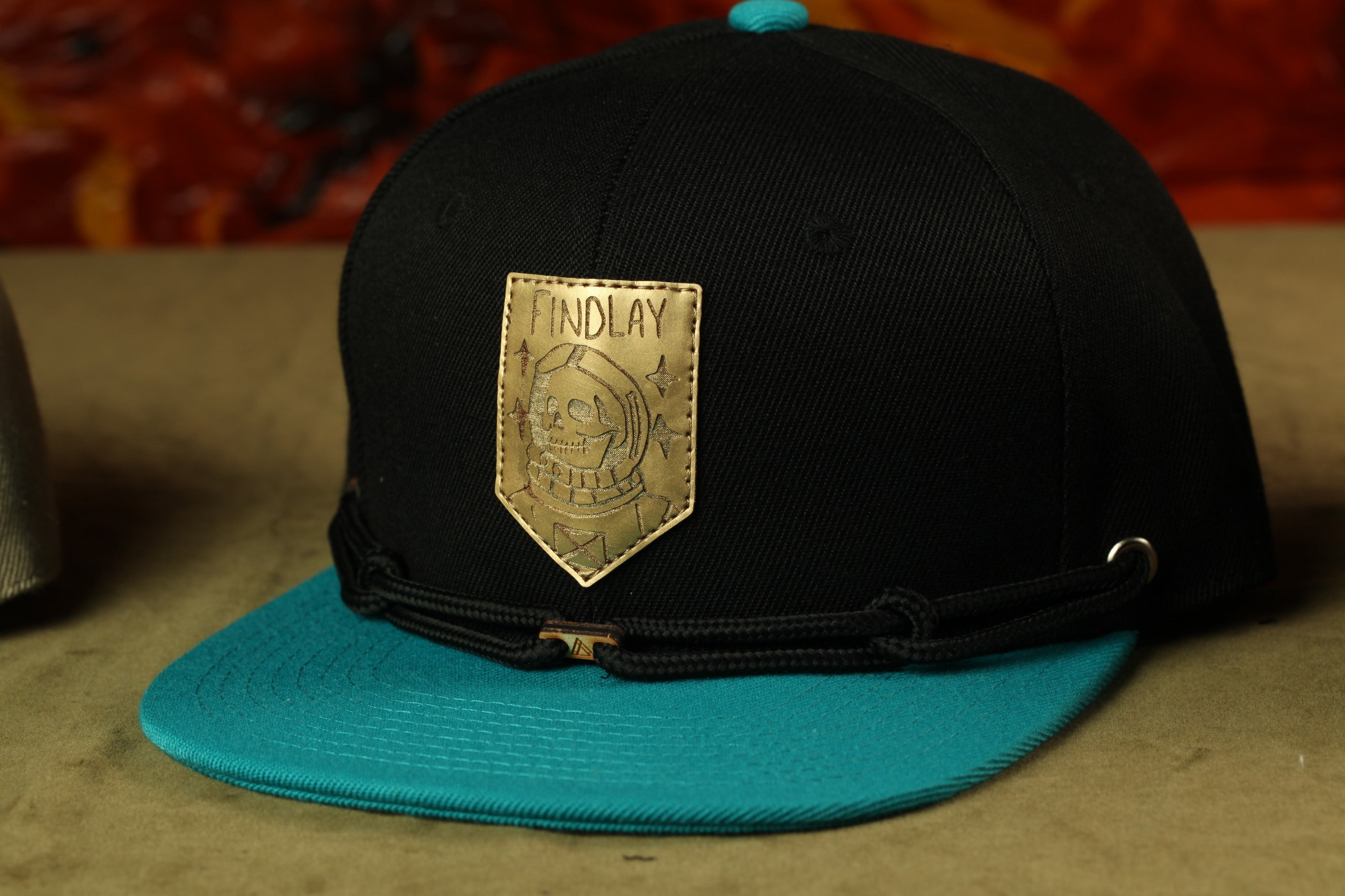 A black baseball cap with a gold patch on the front, featuring a skull design and the word "INDLAY".