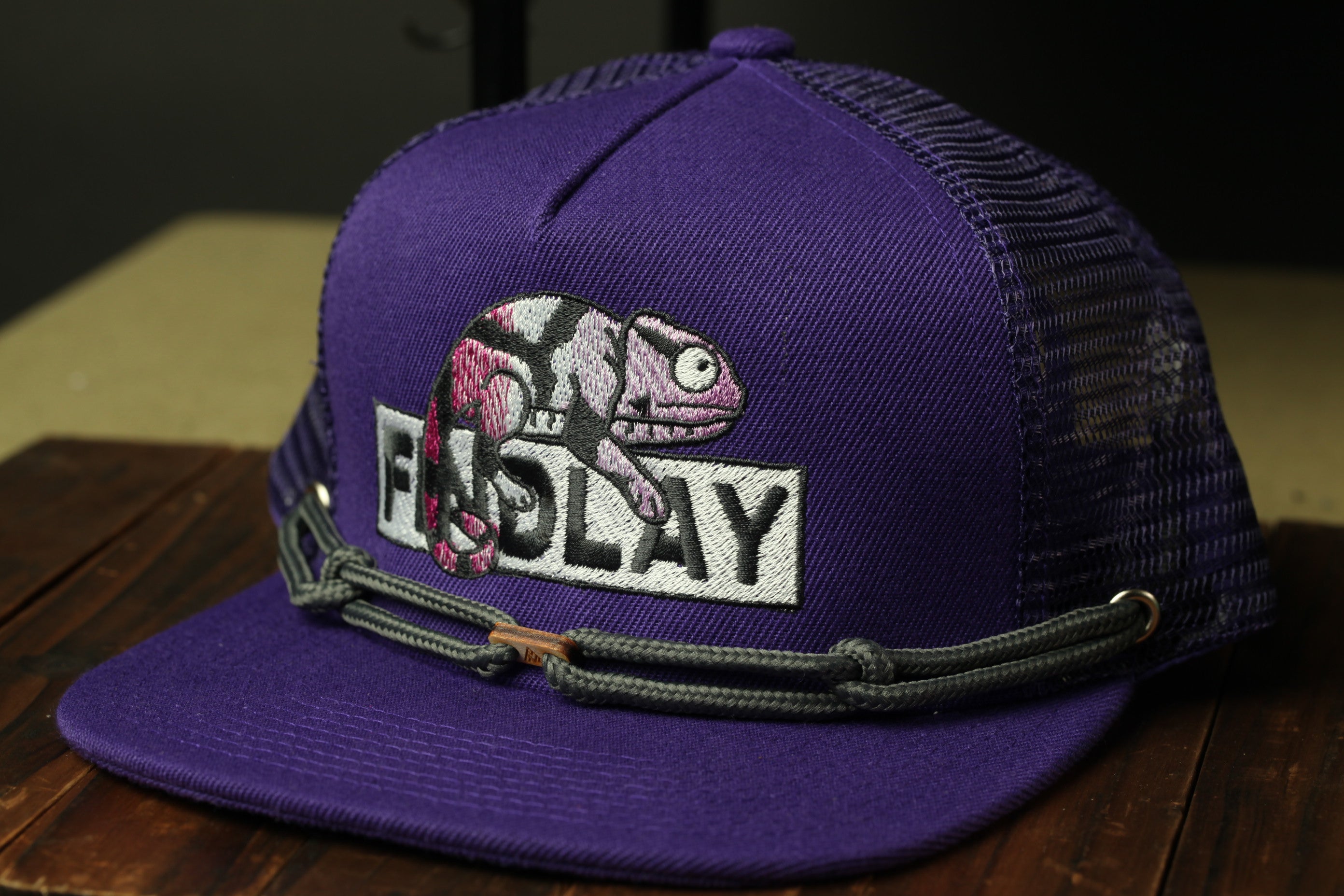 A purple baseball cap with a graphic of a chameleon on it, sitting on a wooden surface.
