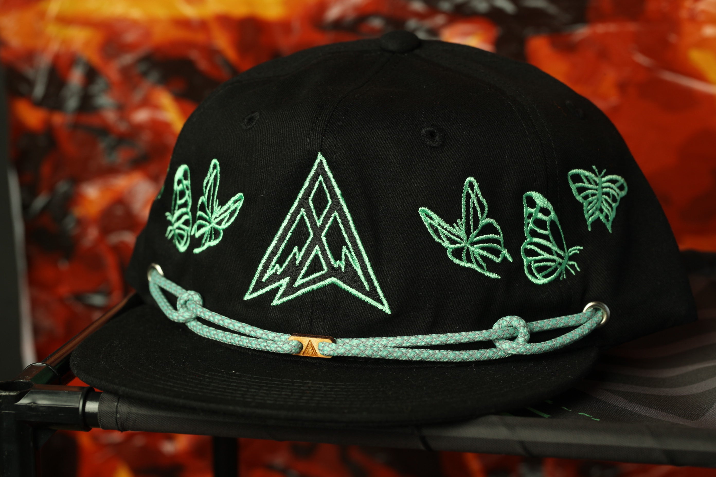A black baseball cap with a green geometric design and butterflies embroidered on it, displayed against a red and orange background.
