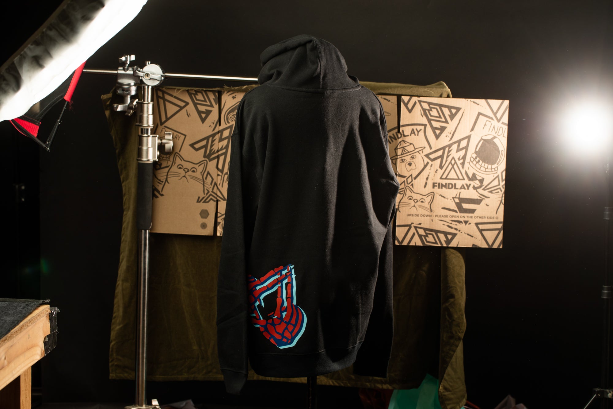 A black hoodie with a graphic design on the back is displayed in a dark room, with cardboard boxes and a light source in the background.