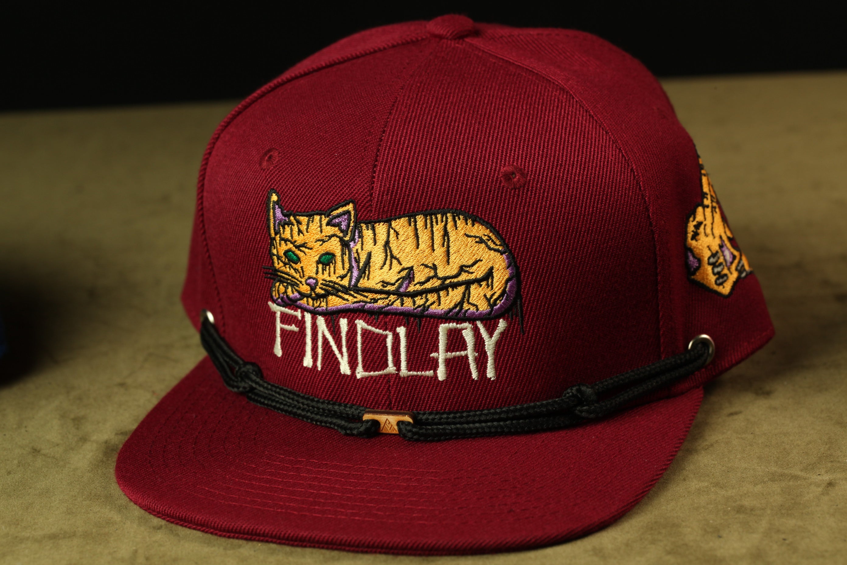 A maroon baseball cap with a cat embroidered on it, featuring the word "FINDLAY" printed on it.